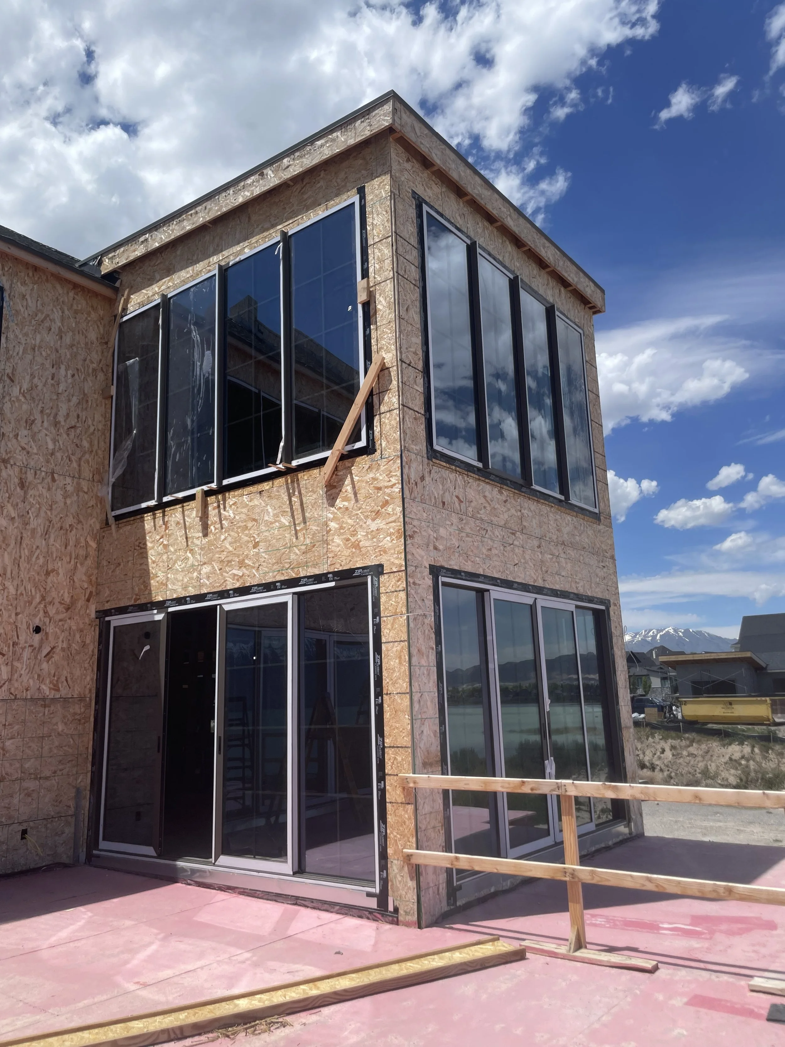 Building under construction with large glass windows, plywood siding, and a pink concrete base, overlooking snow-capped mountains and a blue sky with clouds.