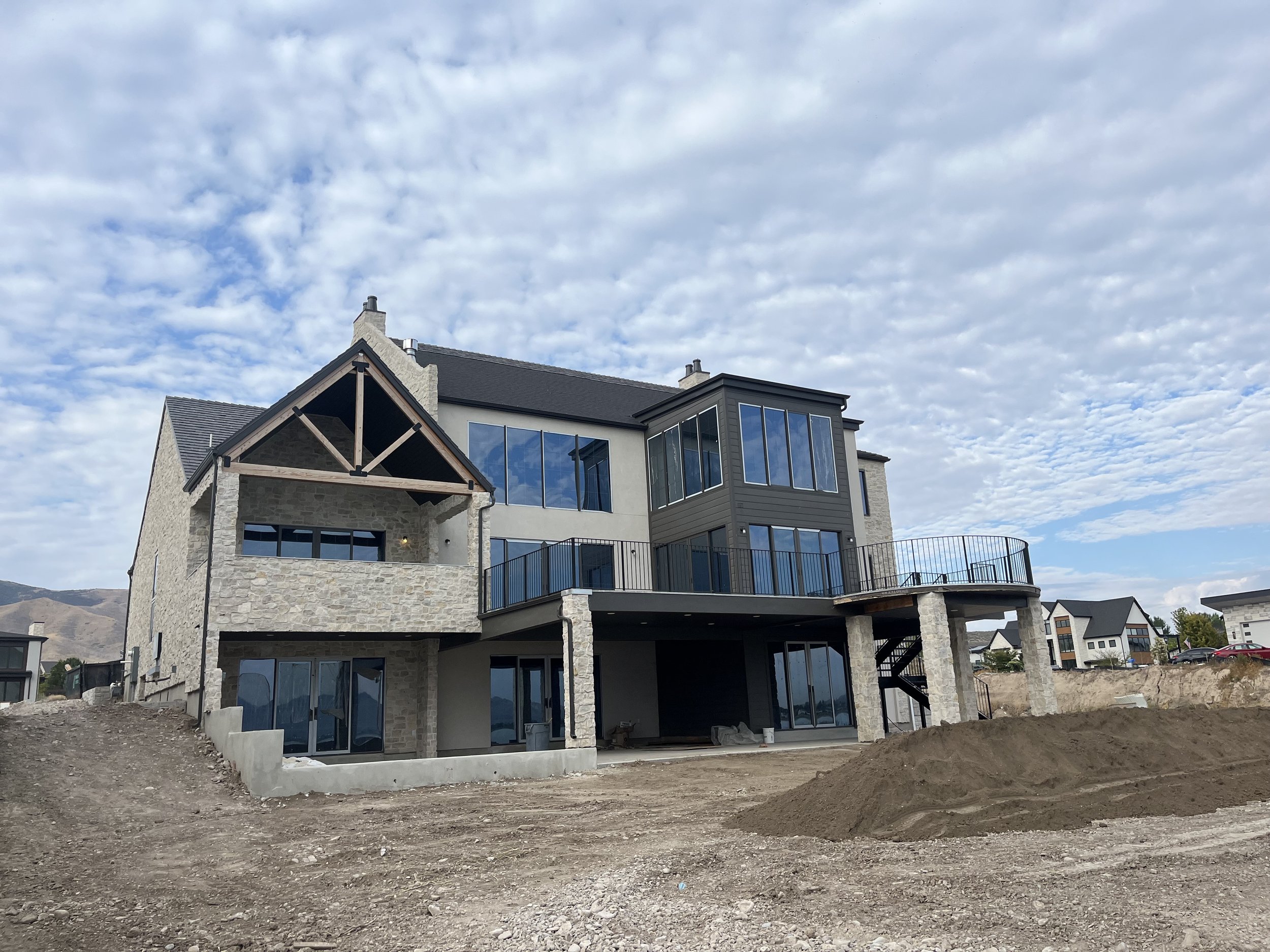 A large modern house under construction with stone and siding exterior, large glass windows, and multiple balconies, situated in a mountainous area under a partly cloudy sky.