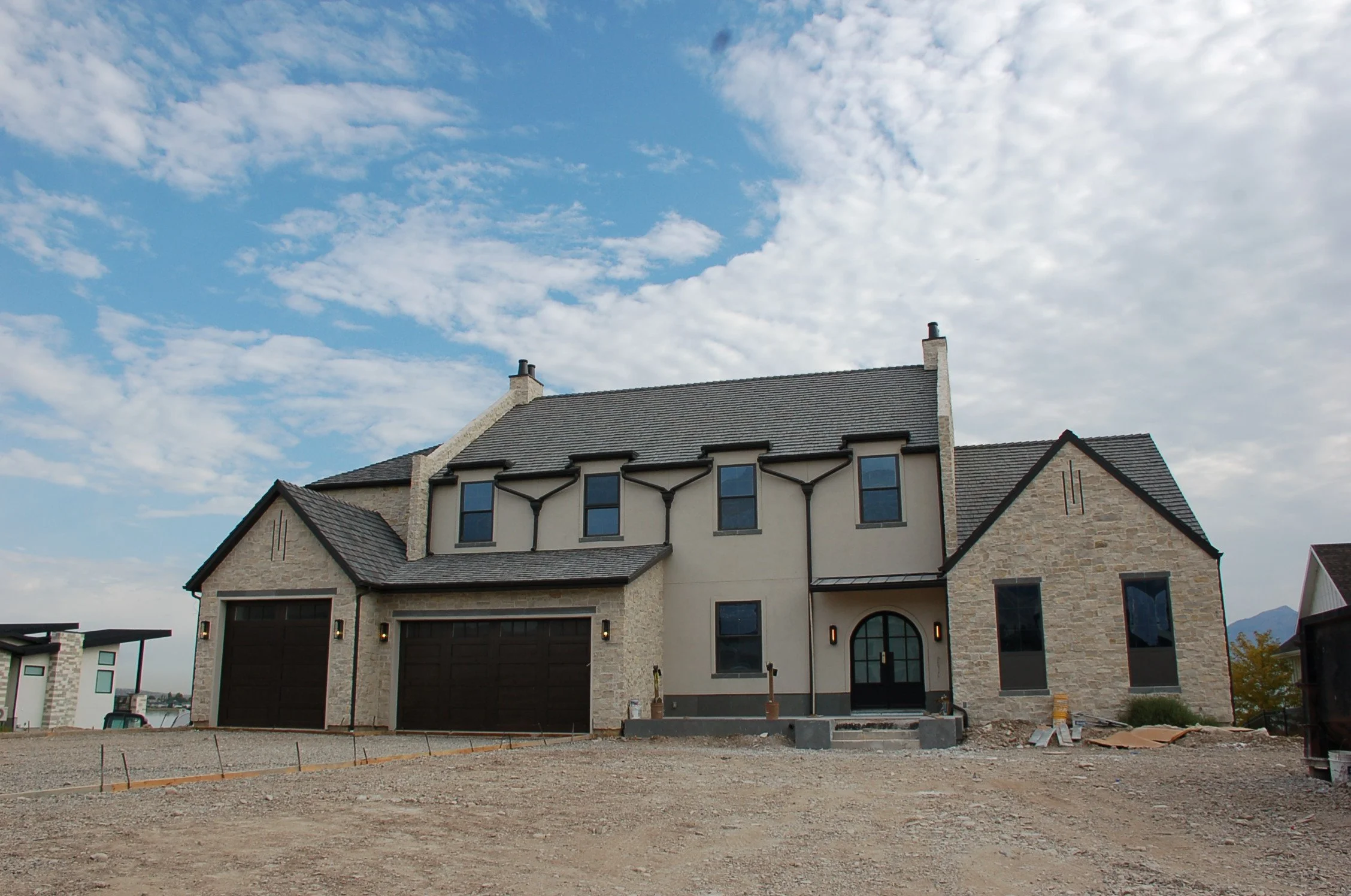 A newly built house with a dark garage door, a black front door with an arched window, and stone and beige siding. The house has five windows on the top floor, some with black trim. The front yard is dirt and gravel, with construction materials nearb