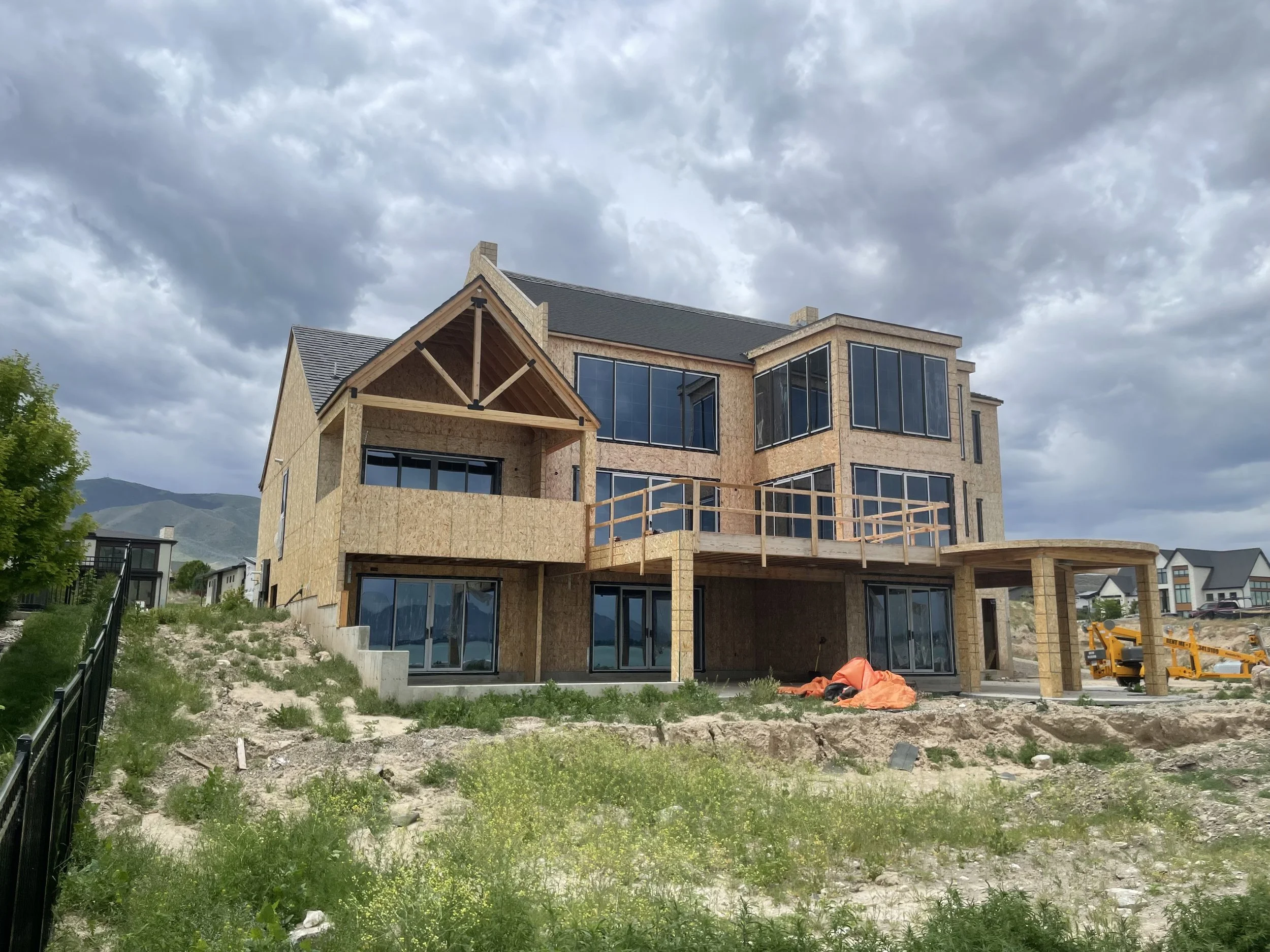 A multi-story house under construction with a wooden frame, large glass windows, and a partially completed deck. The house is set in a semi-rural area with mountains in the background, cloudy sky overhead, and some construction equipment and material