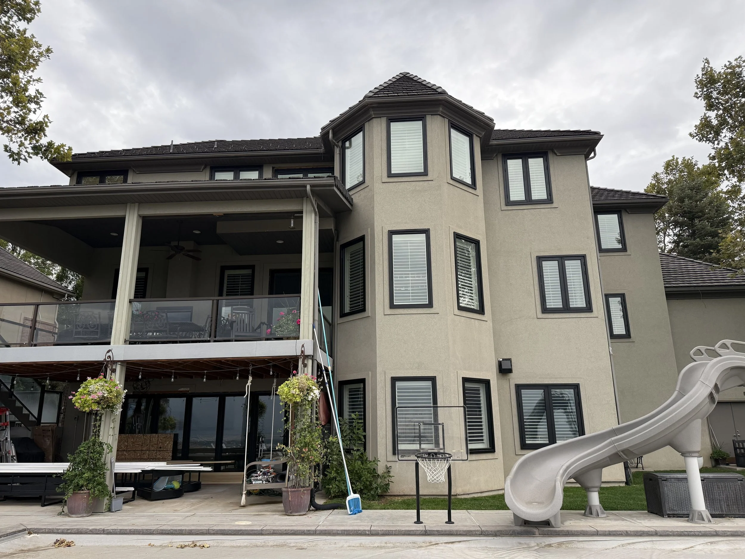 Multi-story house with a beige exterior and multiple black-trimmed windows, a covered patio with hanging plants and patio furniture, a basketball hoop, and a large outdoor slide, under a cloudy sky.