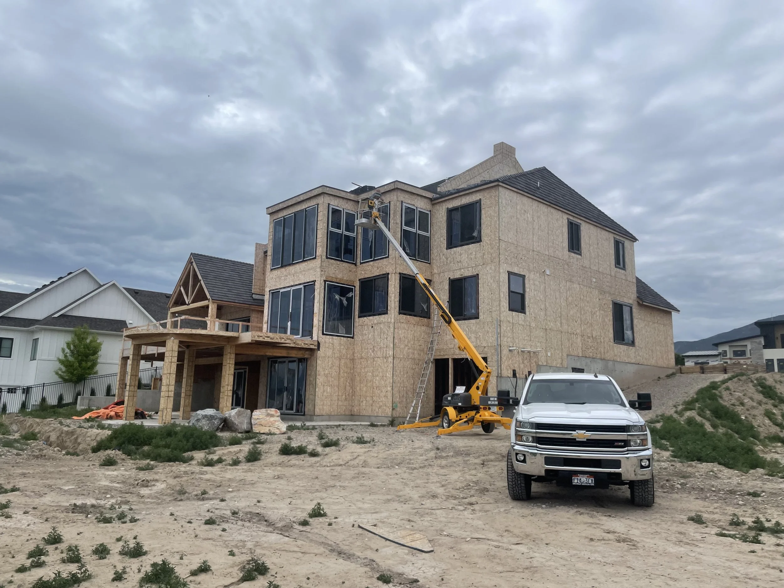 A multi-story house under construction with exposed wood framing and large windows, a yellow construction lift, a white pickup truck, and a cloudy sky in the background.