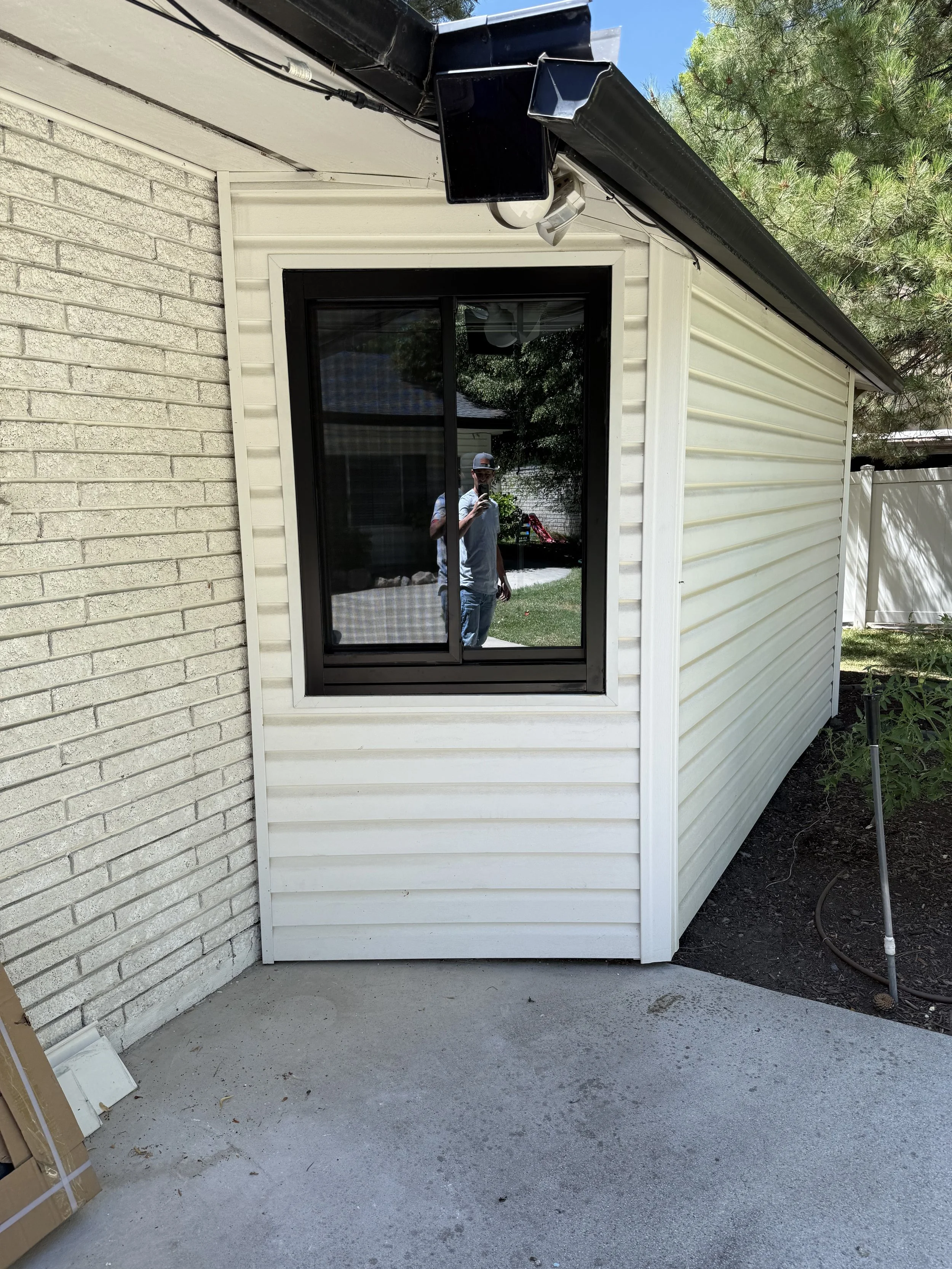 A house exterior showing a window with black frame and glass panes reflecting a person taking a photo, with a backyard visible in the reflection. The house has white vinyl siding, and there is a black gutter system on the roof. The concrete patio is 