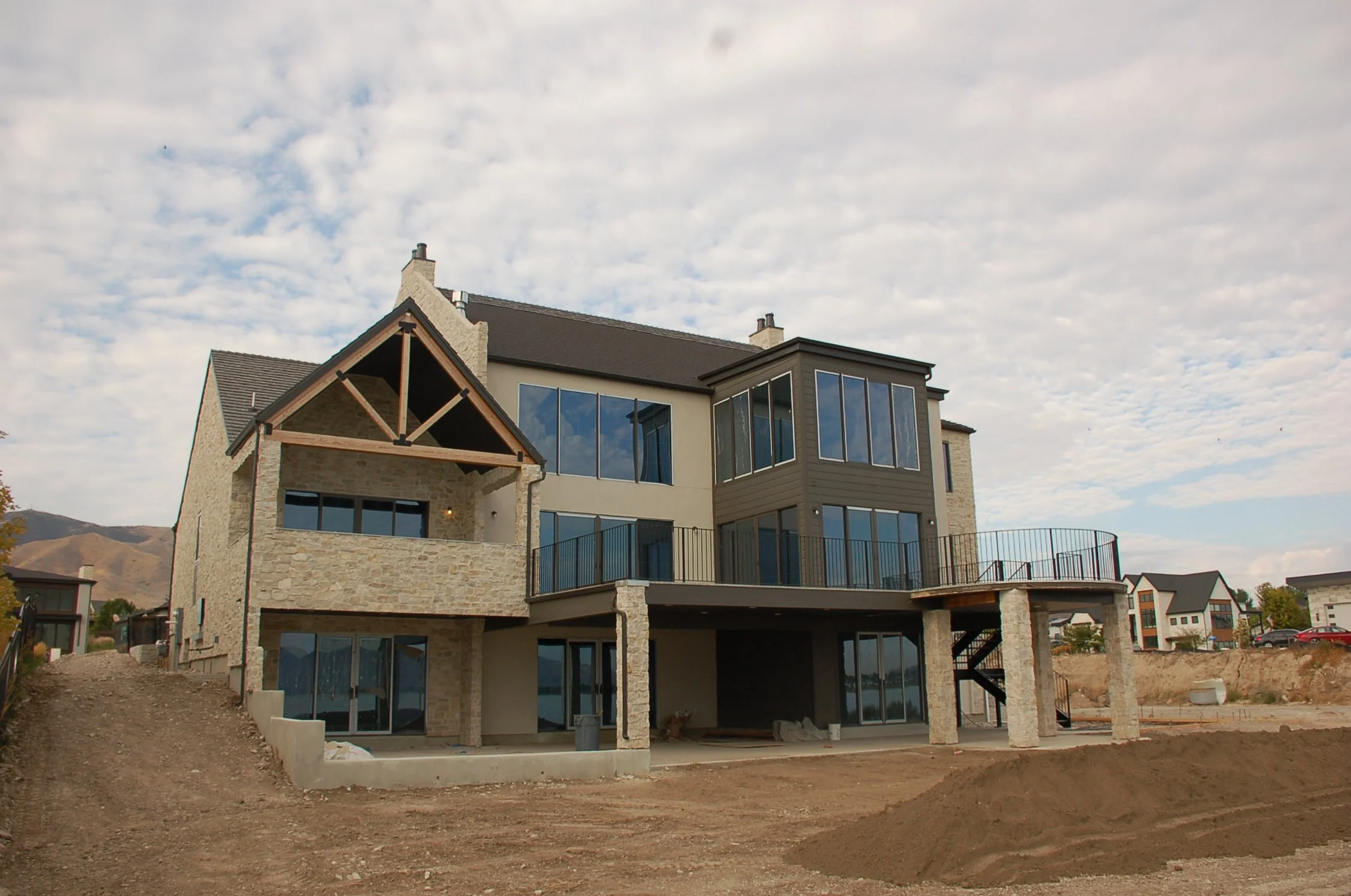 A large modern house under construction with stone and siding exterior, multiple large windows, a balcony, and stairs leading to the ground. The yard is unfinished with dirt and construction materials.