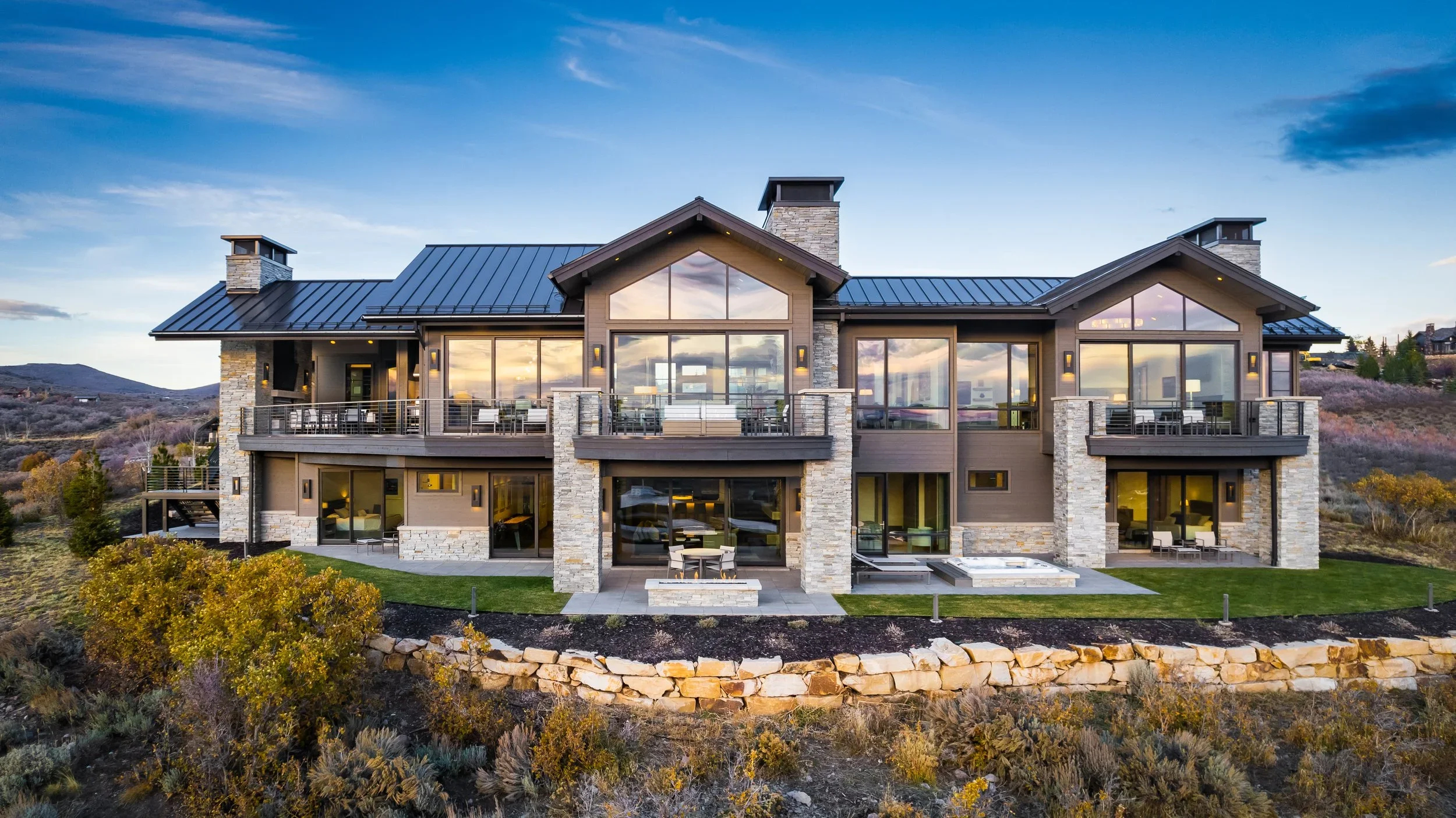 Large modern house with multiple balconies, glass walls, stone accents, and a backyard yard with a small fire pit and outdoor furniture, set against a scenic landscape at sunset