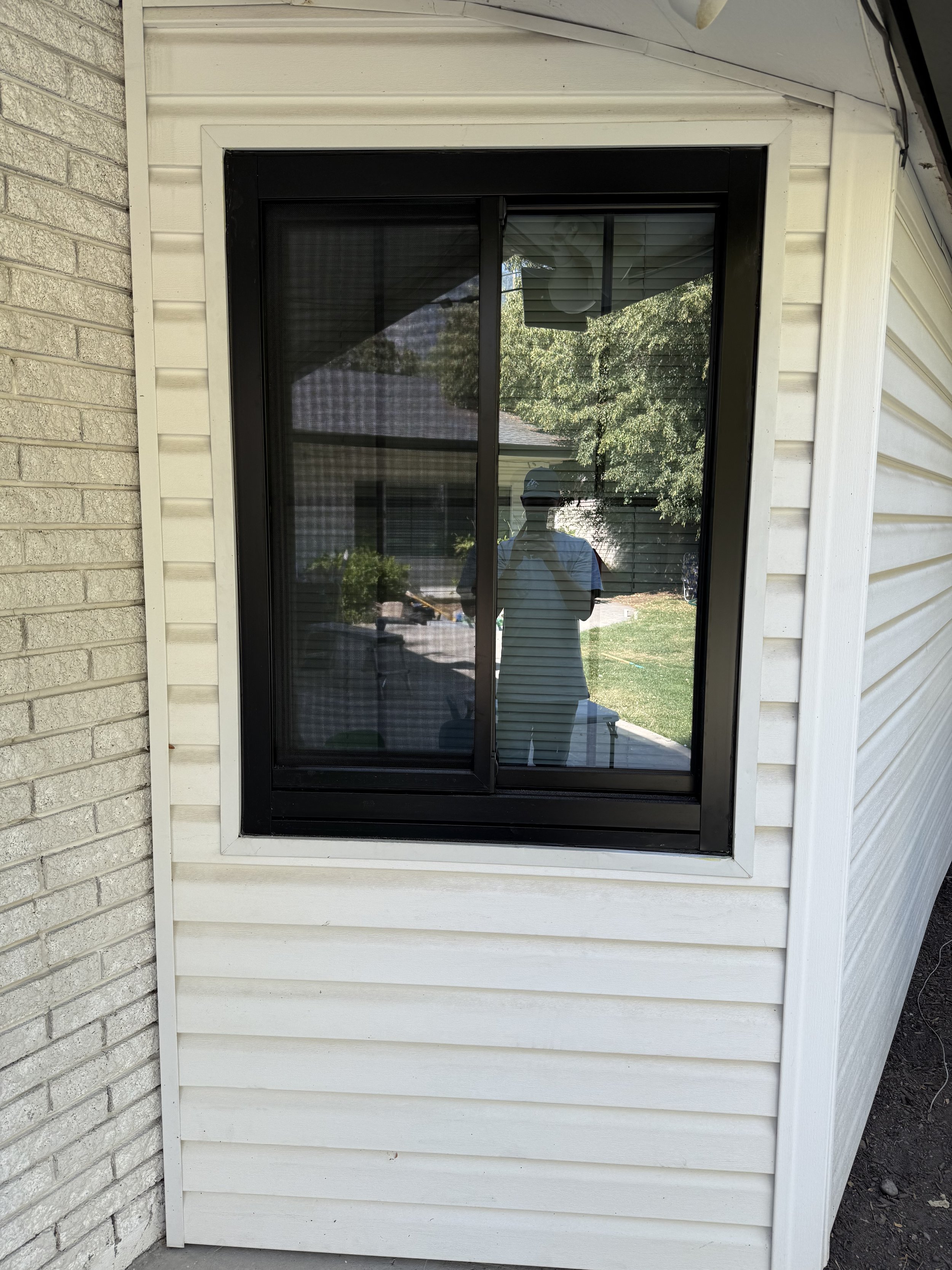 Exterior view of a window with a screen, reflecting the person taking the photo and the surrounding yard.