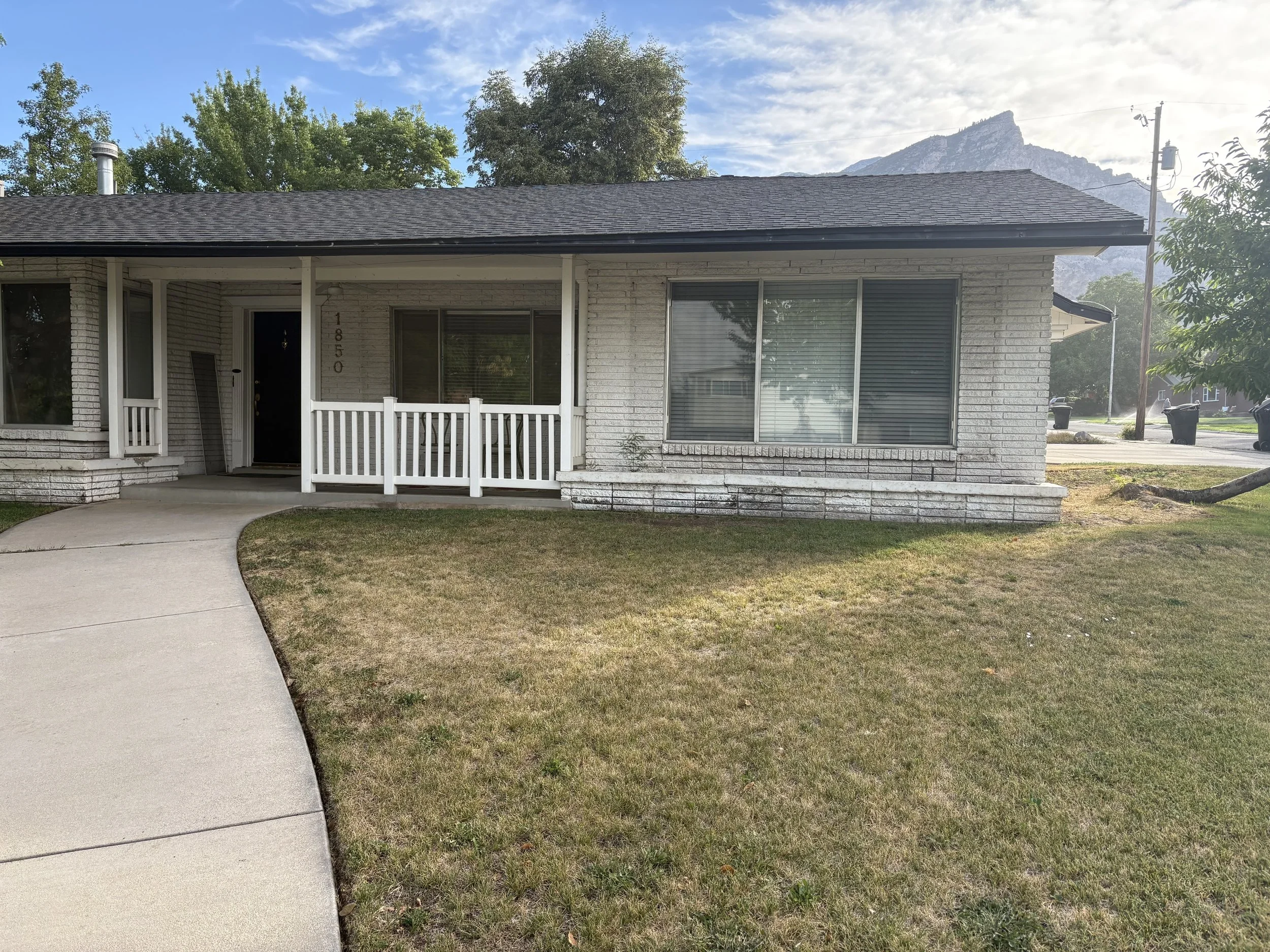 A single-story house with a white brick exterior and a black roof. It has a small front porch with white railings, a black door, and large windows. The front yard has a curved concrete walkway and a grassy lawn, with some trees and mountains in the b