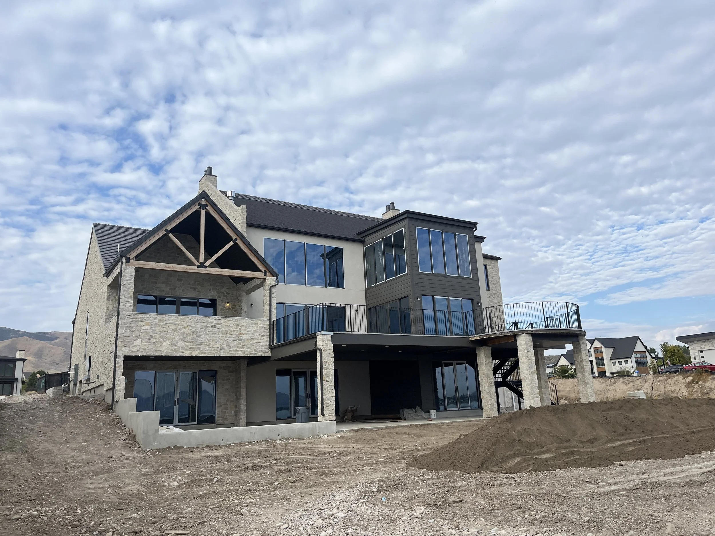 A large, multi-story house under construction with stone and gray siding, large glass windows, and a curved balcony. The ground in front of the house is bare dirt, with some construction materials nearby. The sky is partly cloudy.