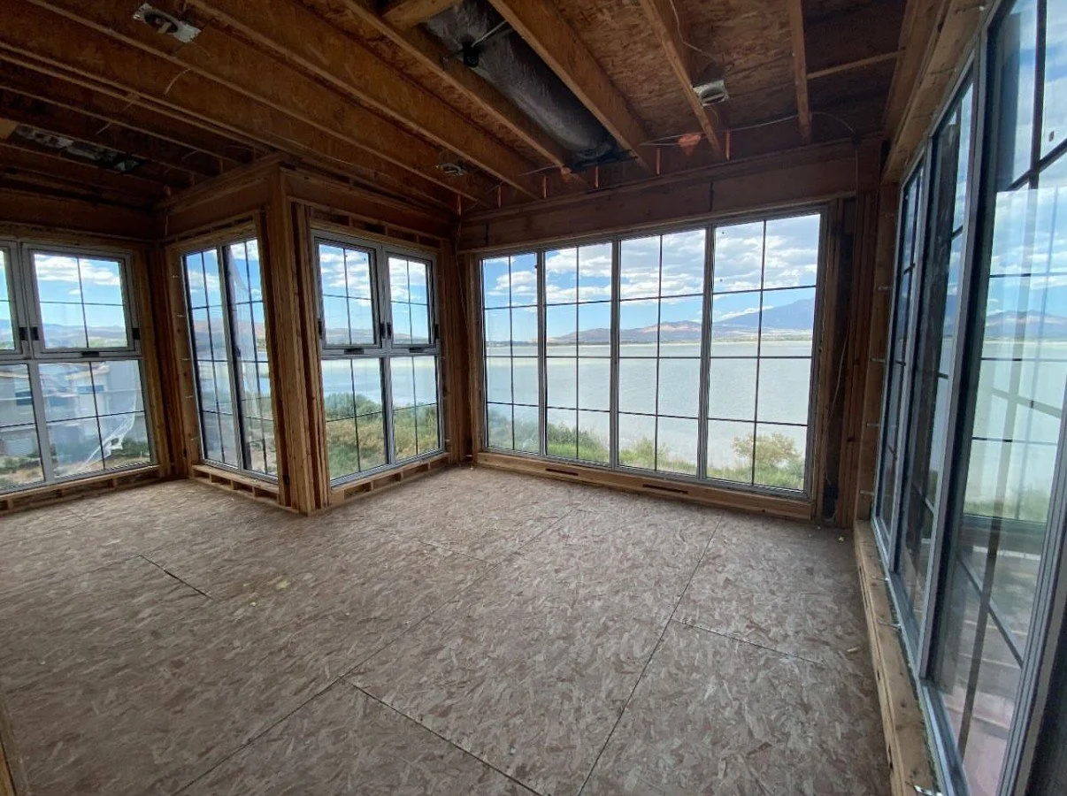 Unfinished room with large windows overlooking a body of water and mountains in the distance. The room has exposed wooden beams and OSB flooring, and is under construction.