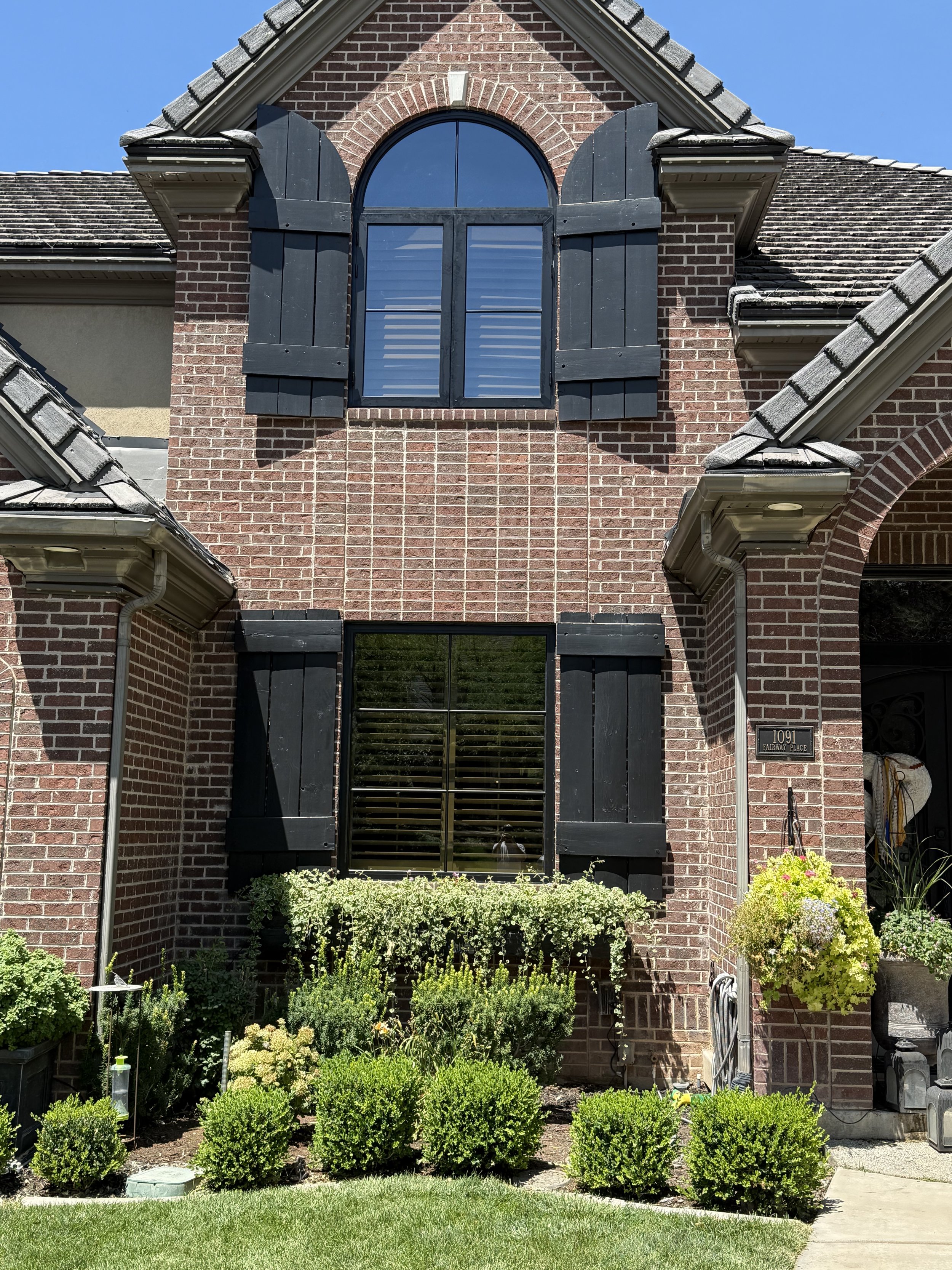 Front view of a brick house with black shutters, an arched window on the upper level, and a front yard with green bushes and potted plants.