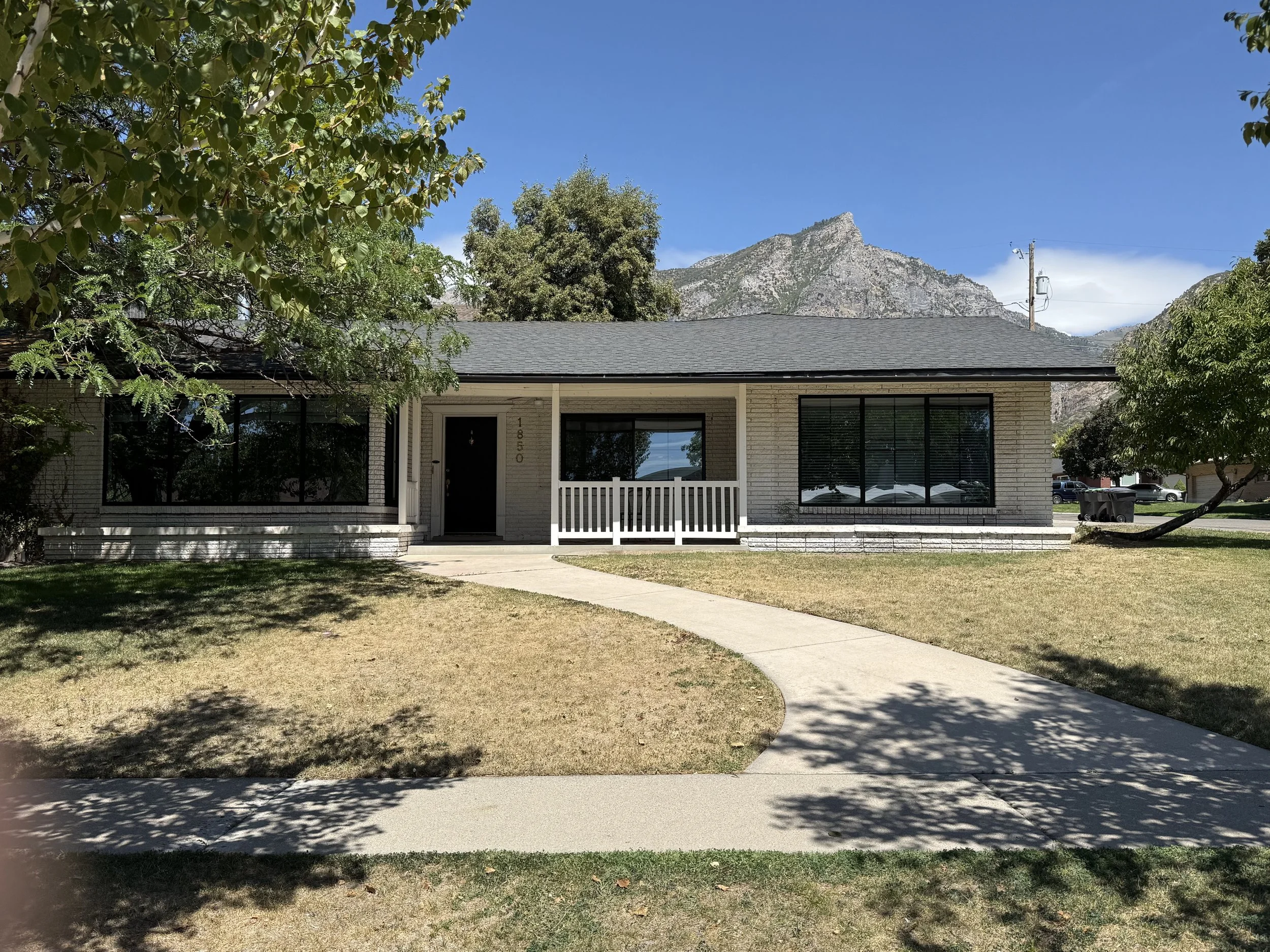 A single-story house with white brick exterior and black front door, surrounded by a lawn with trees and mountains in the background