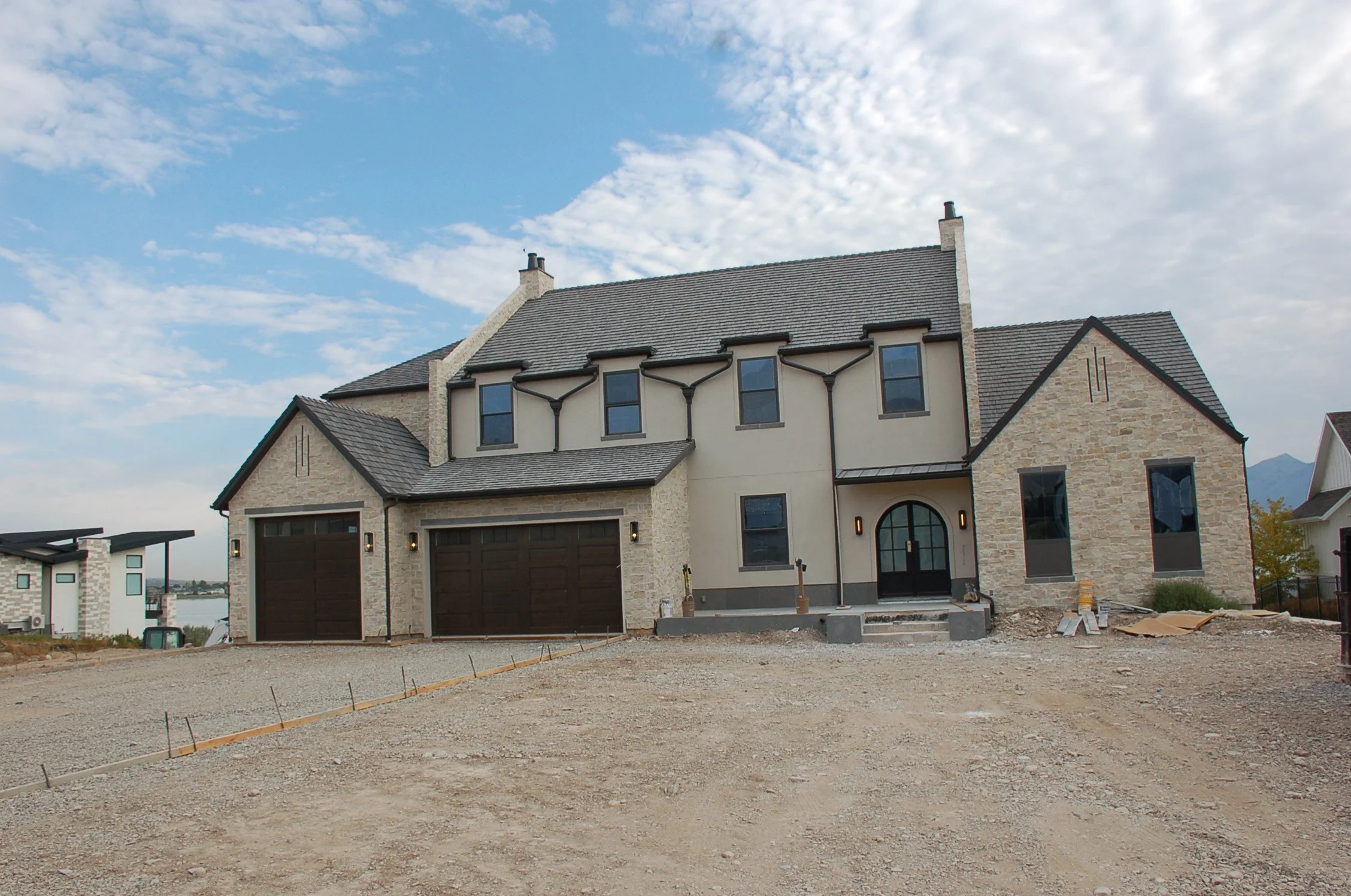 Newly built house with dark wood garage doors, stone and stucco exterior, arched front door, and a dirt yard.
