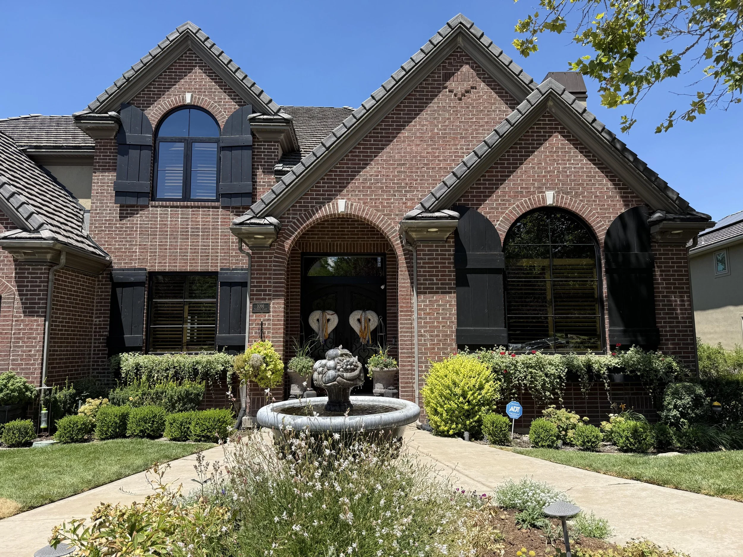 A brick house with black shutters, arched and rectangular windows, a small front yard with a fountain, and well-maintained garden plants.