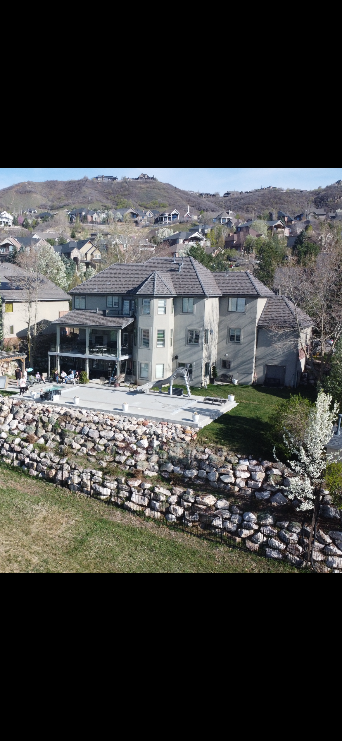 A large multi-story house with a backyard patio and a small slide, surrounded by a stone retaining wall and a grassy area. The house is located in a hilly residential neighborhood with other houses on the hills in the background.