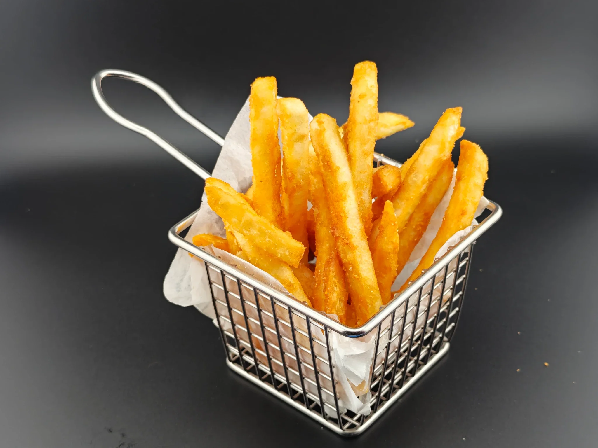 A small metal basket filled with golden, crispy French fries lined with white paper, set against a dark background.