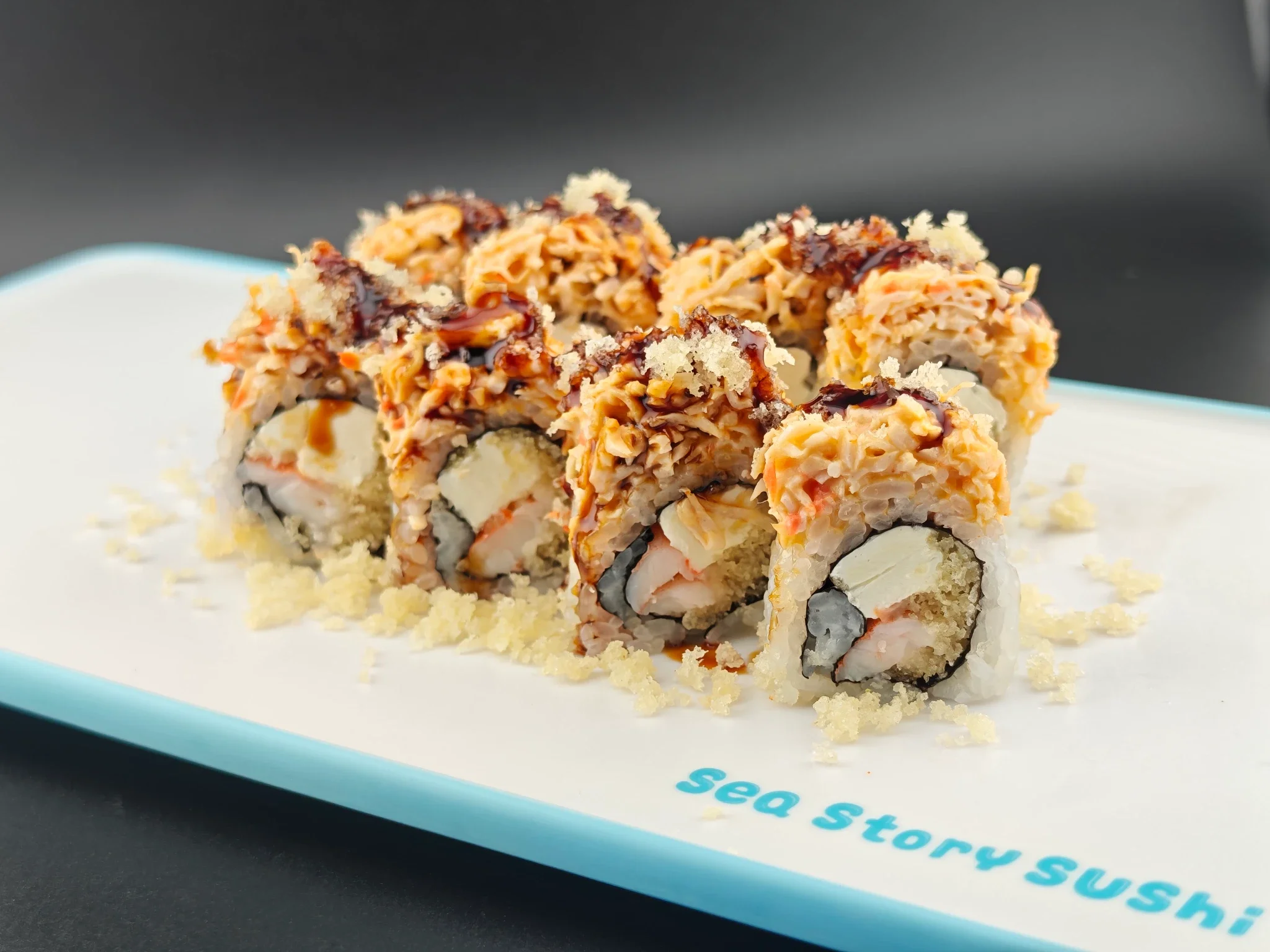 Close-up of sushi rolls topped with crunchy flakes, drizzled with sauce, and served on a white plate with a blue edge.