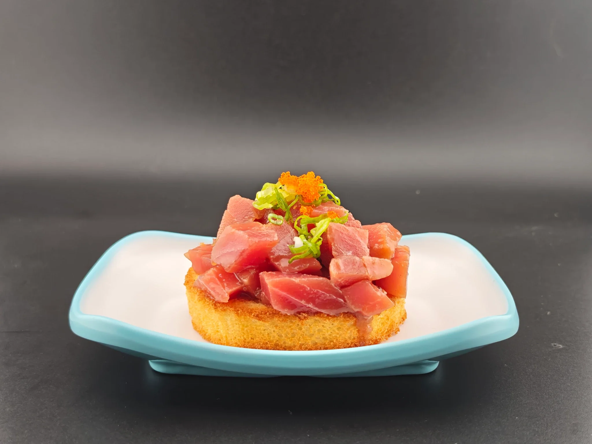 Close-up of a piece of toast topped with raw tuna, chopped green onions, and orange fish roe, served on a blue dish against a dark background.