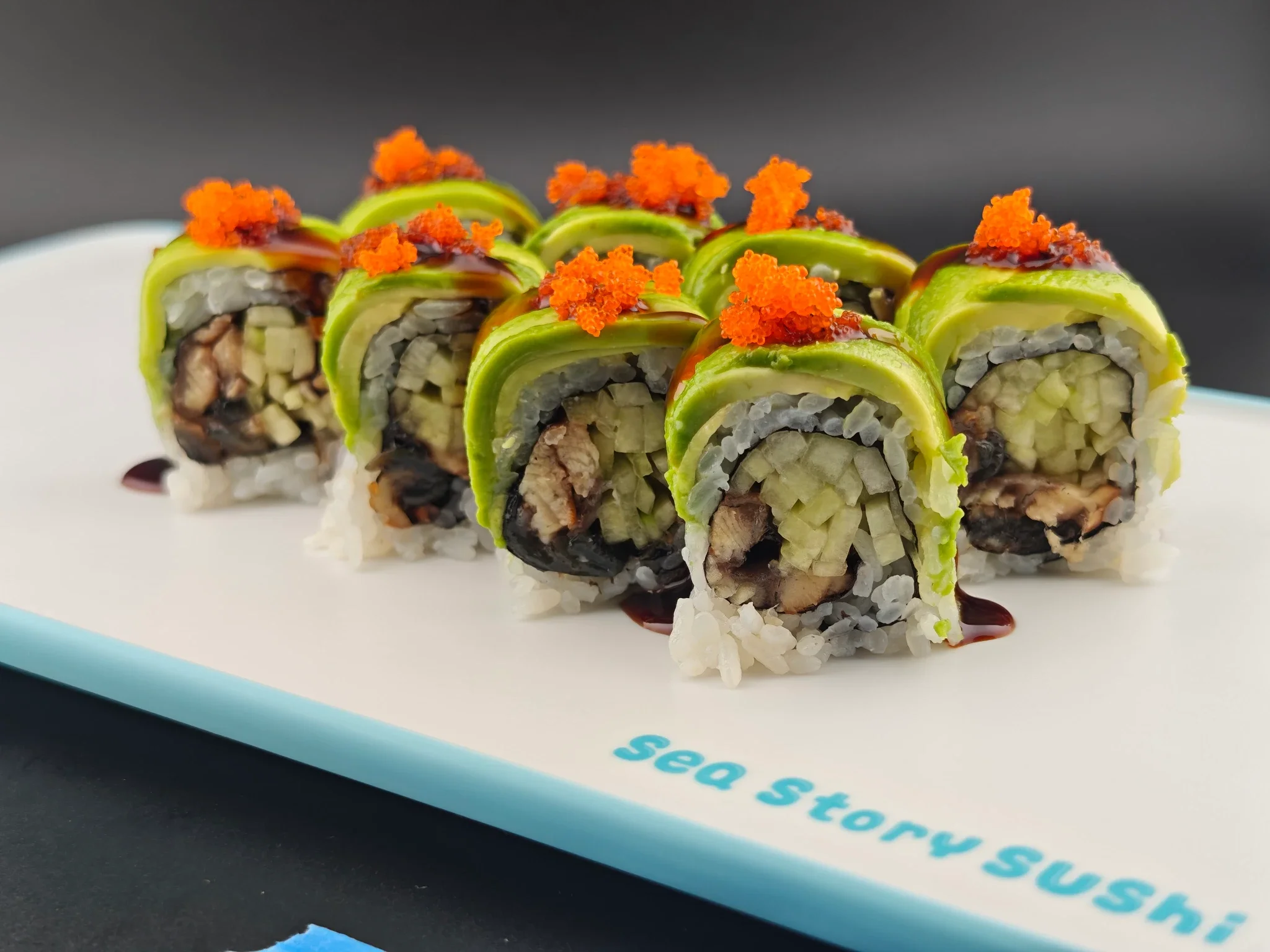 Close-up of a sushi roll topped with orange fish roe, drizzled with dark sauce, on a white plate with blue text.