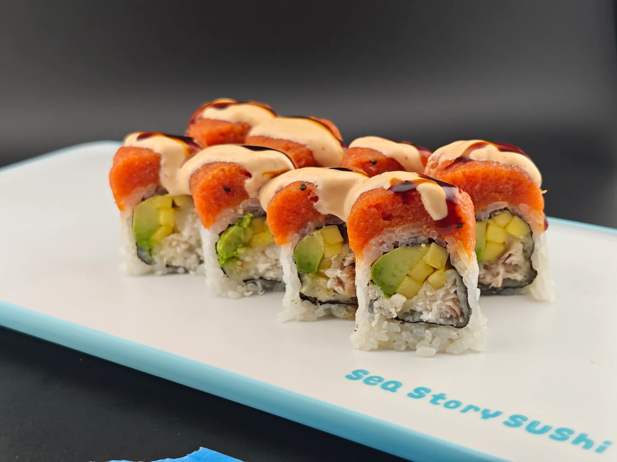 Close-up of sliced sushi roll with avocado, crab, and sauce on top, served on a light blue plate against a dark background.