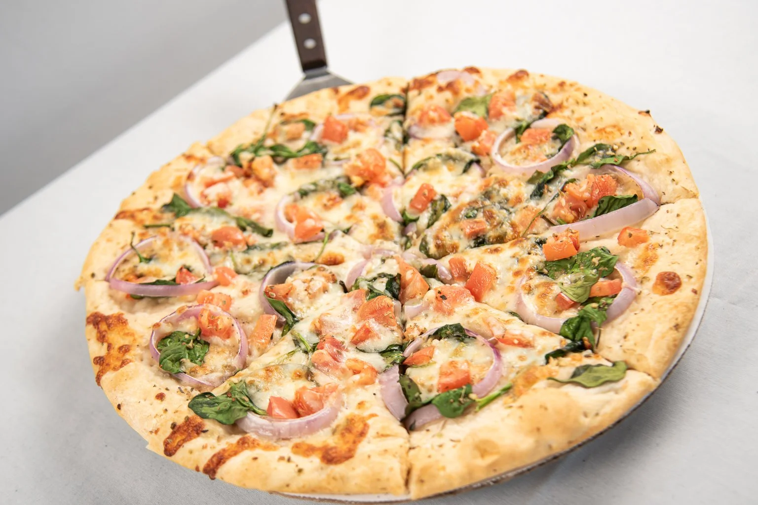 A freshly baked pizza with cheese, tomato, spinach, and onions on a metal pizza paddle.