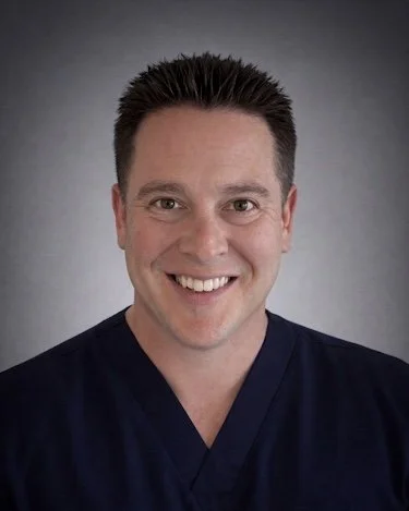 A smiling man with dark hair, wearing a navy blazer, a light blue and pink plaid shirt, and a dark blue tie, standing outside near a beige wall and a closed gray door.