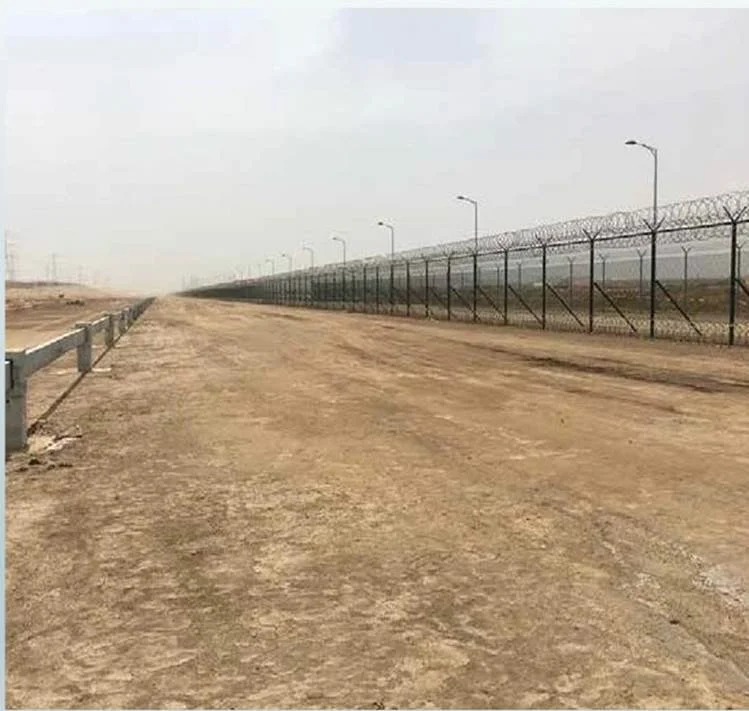 Empty dirt road next to a tall fence topped with barbed wire, with streetlights along the fence in a hazy, barren landscape.