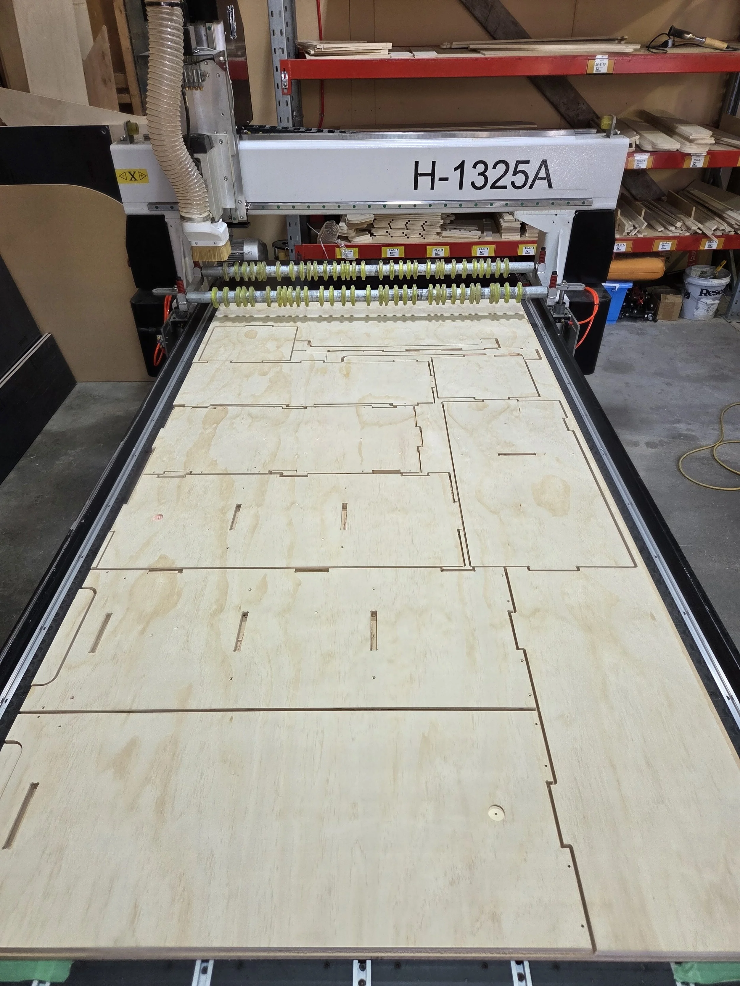 A CNC machine cutting wood panels with outlined shapes. The machine is labeled H-1325A and is in a workshop setting, with shelves holding various wood pieces and tools in the background.