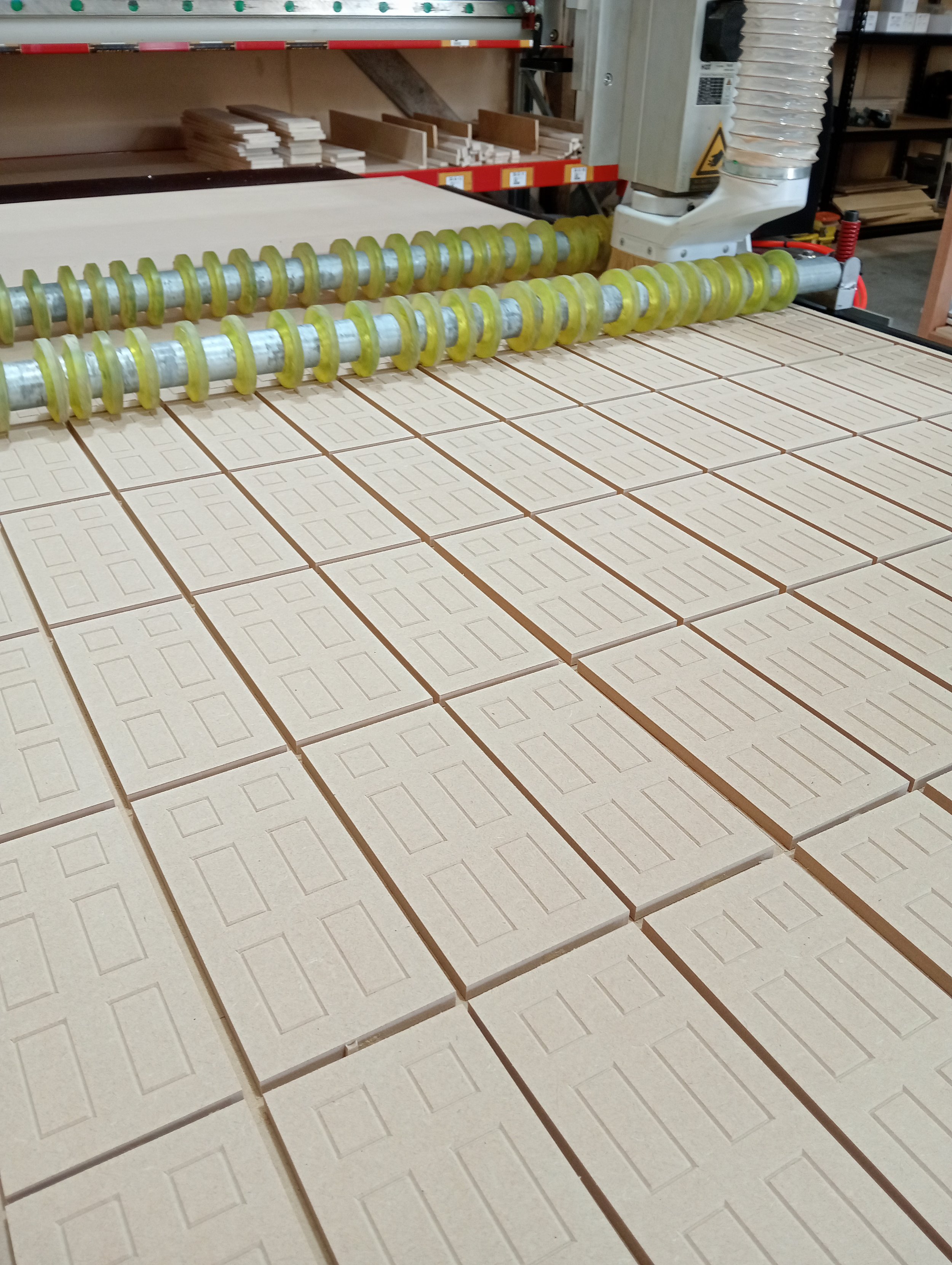 Two large rollers with yellow rings are on a table, with unassembled wooden pieces in front. There are shelves with more wooden pieces and supplies in the background of a woodworking workshop.