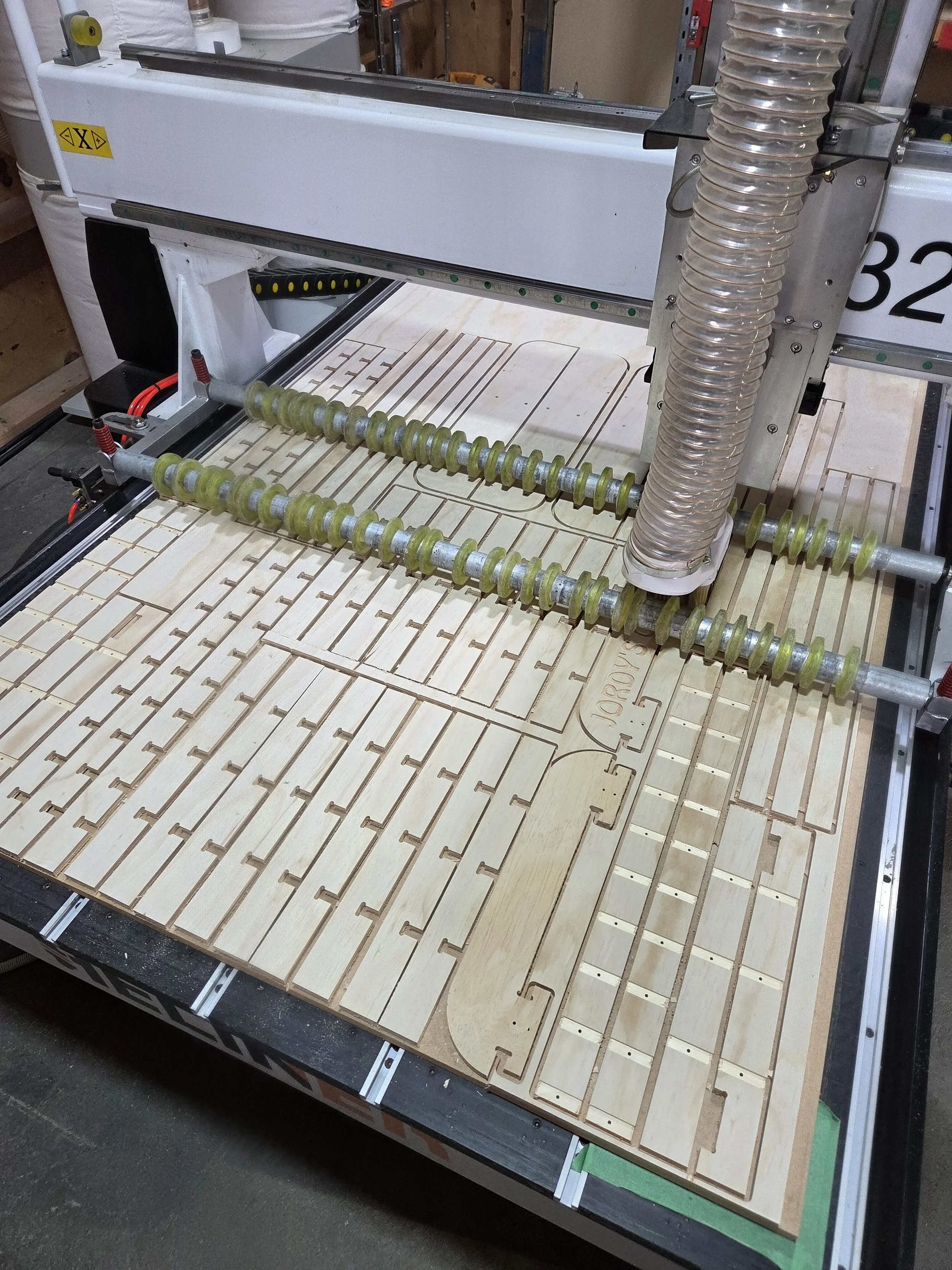 A CNC machine with a wooden project in progress. The machine is working on a wooden sheet with cutouts and slots, with clear flexible tubing attached to the right side of the machine.