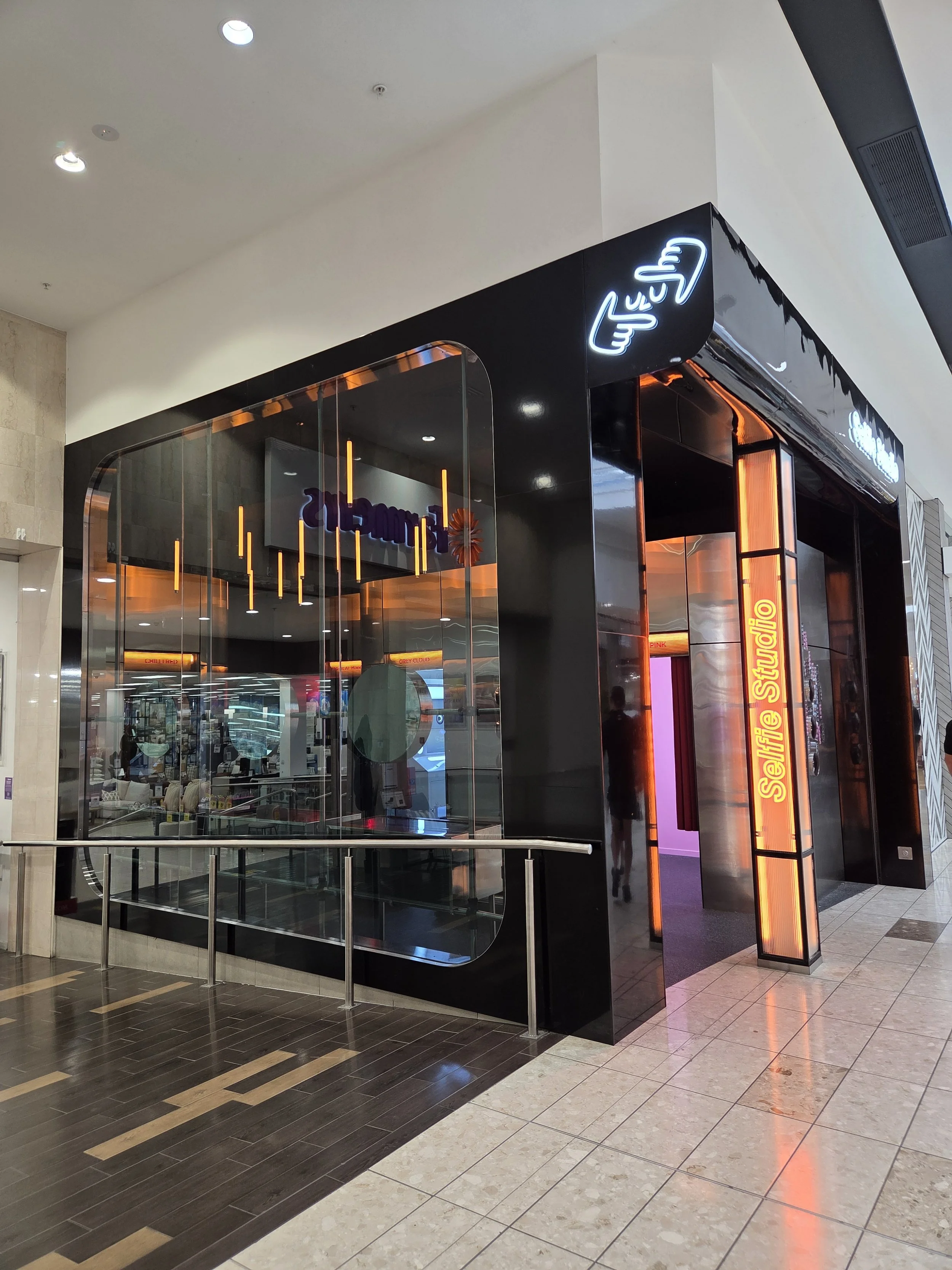 A modern nail salon inside a shopping mall with black, glass, and neon decor. The store features a neon sign with Chinese characters and a sign that says 'Selfie Studio.' The interior includes visible chairs and tables through the glass walls, with a