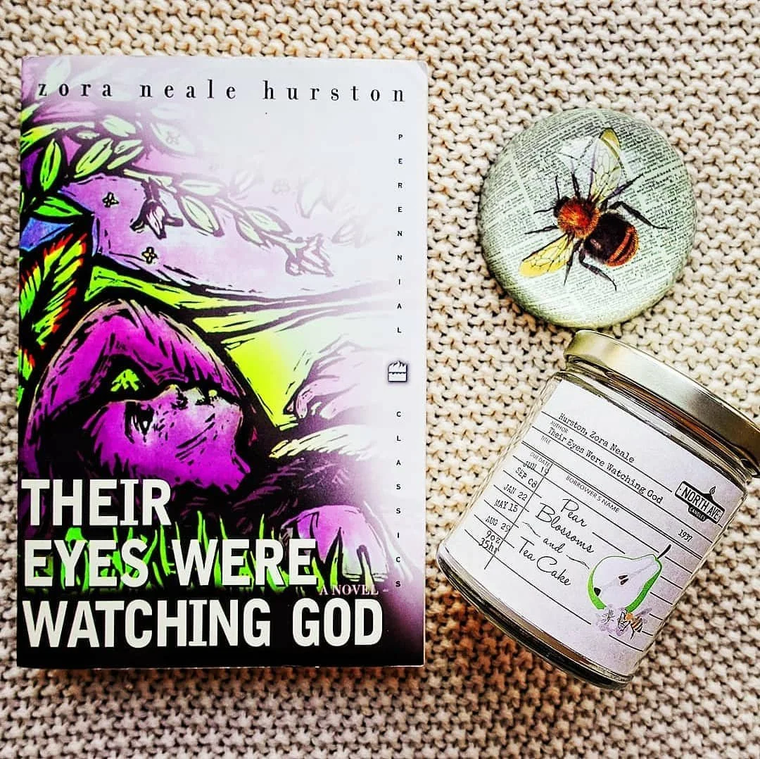 A paperback book titled 'Their Eyes Were Watching God' by Zora Neale Hurston, a decorative pin with a bee illustration, and a glass jar with a label featuring a pear illustration and handwritten notes, all arranged on a textured beige surface.