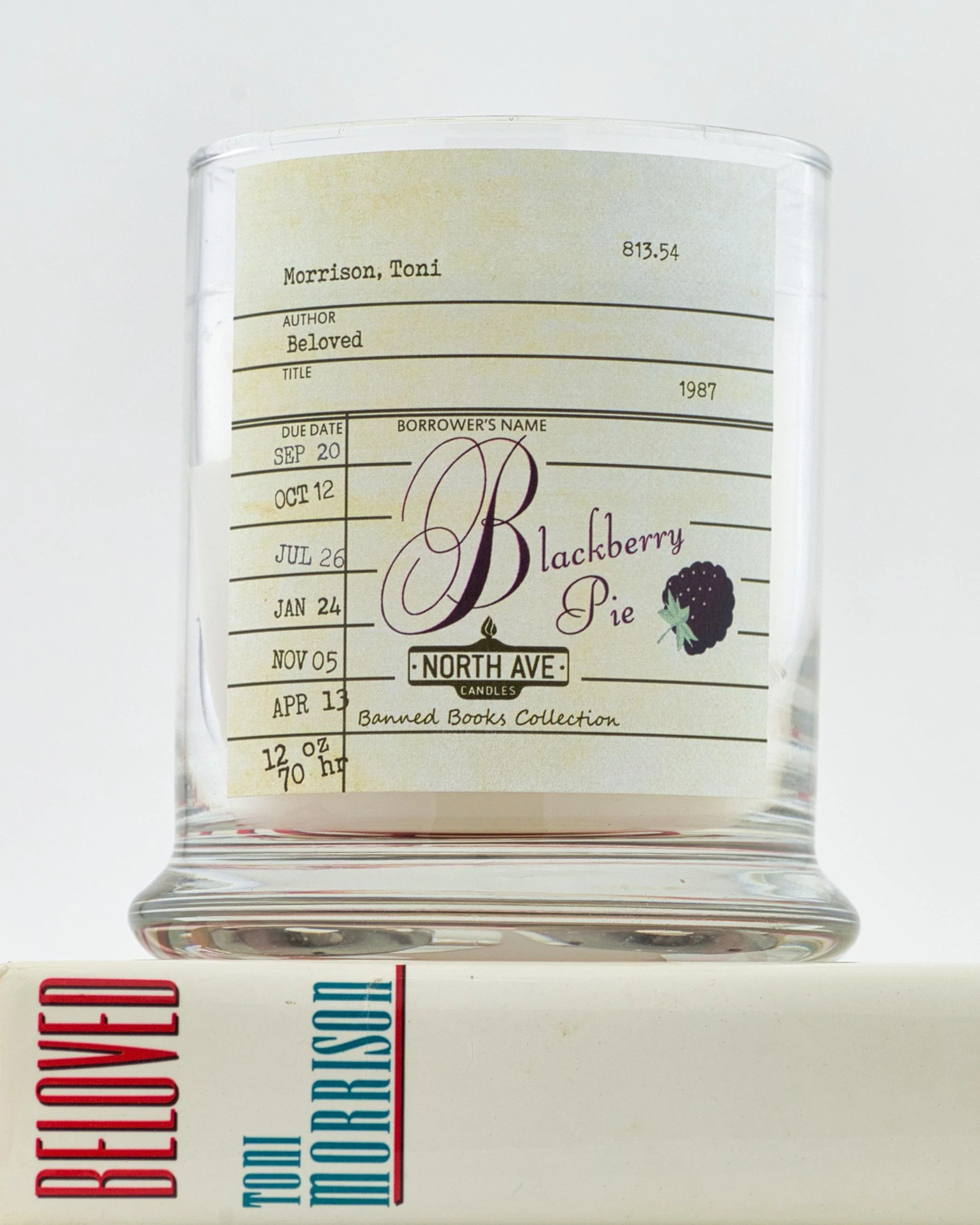 A decorative candle in a clear glass holder with a label resembling a book library card, featuring the title "Blackberry Pie" and a blackberry illustration, placed on top of a box of teal-colored facial tissues.