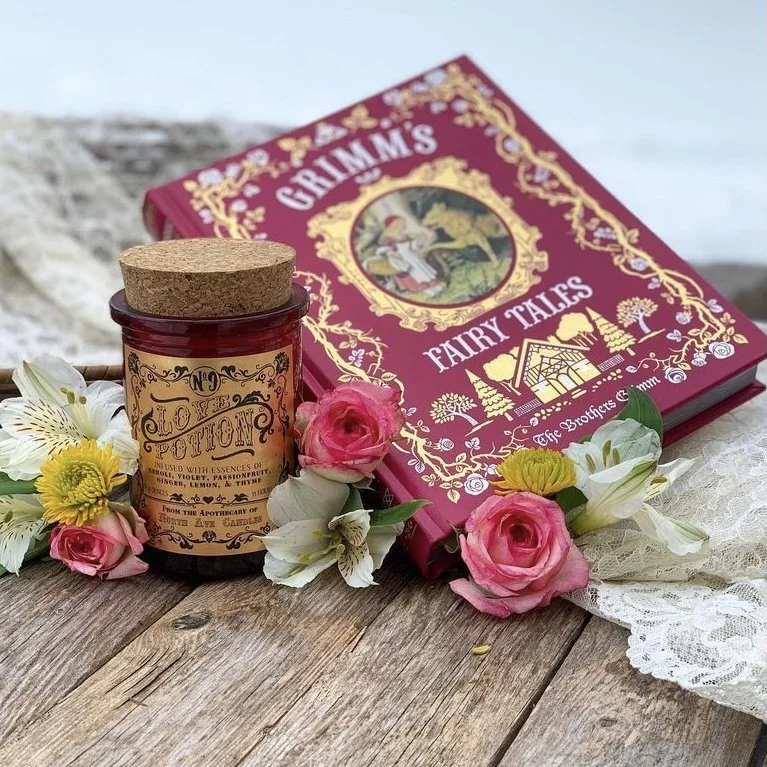A red fairy tale book titled "Grimm's Fairy Tales" lying on a wooden surface surrounded by pink roses, white lilies, and a scented candle with a cork lid.