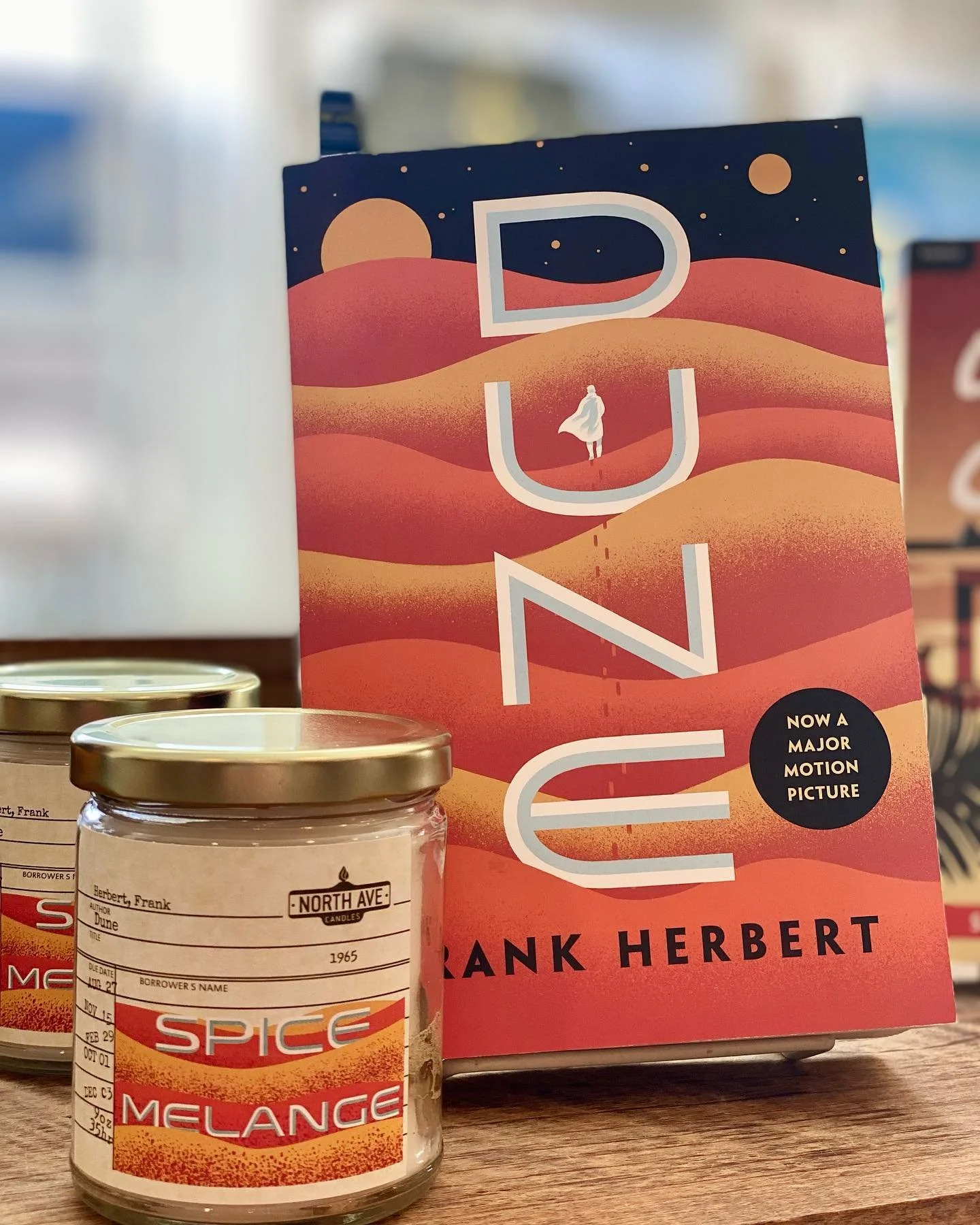 A book by Frank Herbert titled "Dune" with a desert landscape cover featuring rolling sand dunes and a person in a cloak, placed behind candles with labels reading "Herbert, Frank," "Spice" and "Melange."