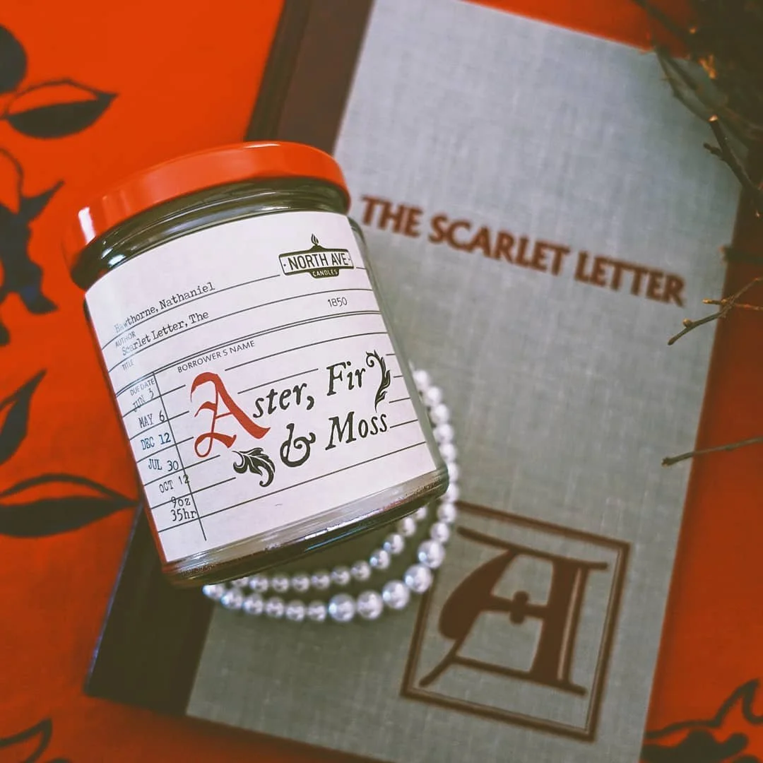 A scented candle in a jar labeled 'Aster, Fir & Moss' from North Ave Candles, placed on a string of pearls. Behind the candle is a book titled 'The Scarlet Letter' and some orange packaging with black leaf illustrations.