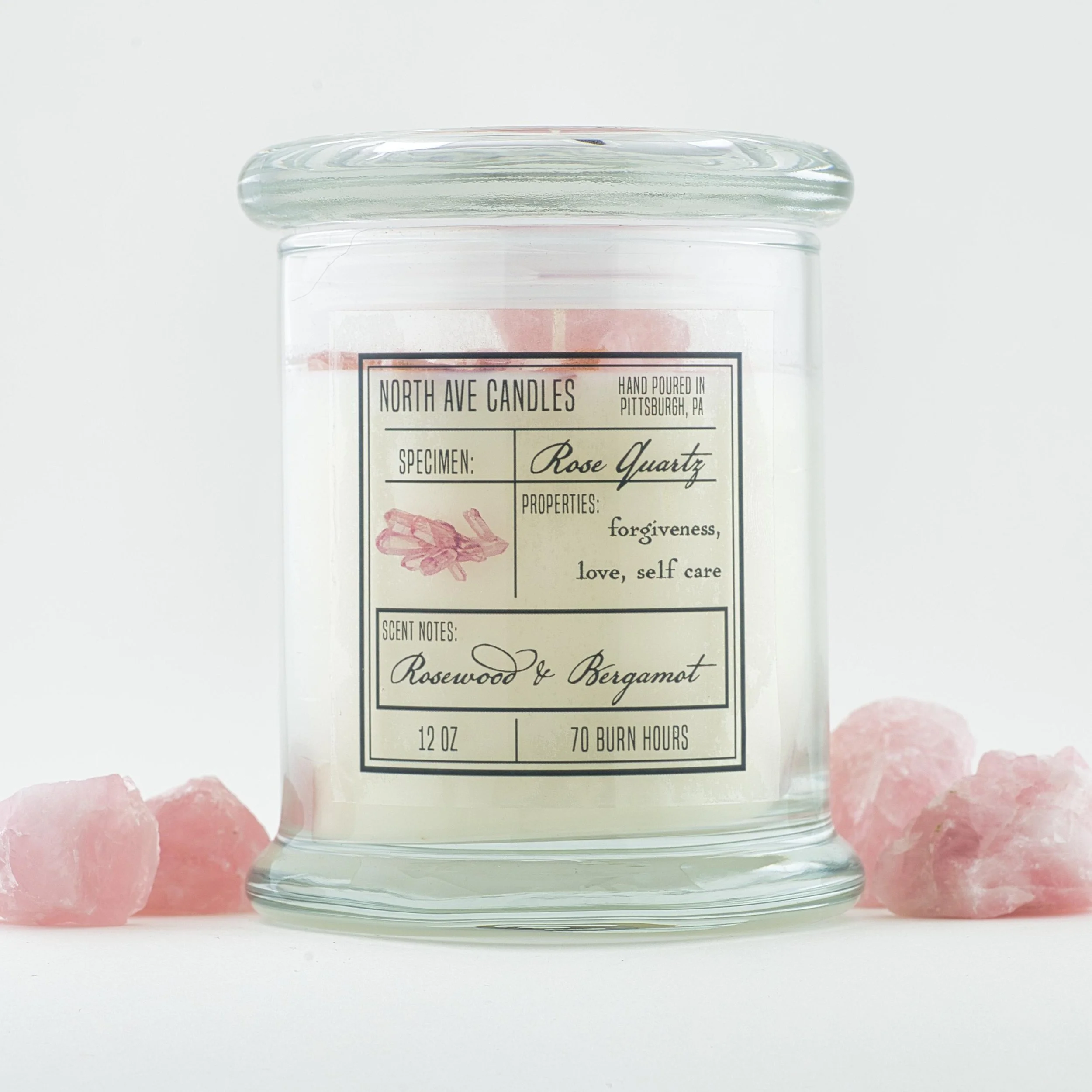 A glass candle jar labeled 'North Ave Candles' with scents of rose quartz and bergamot surrounded by pink salt crystals.