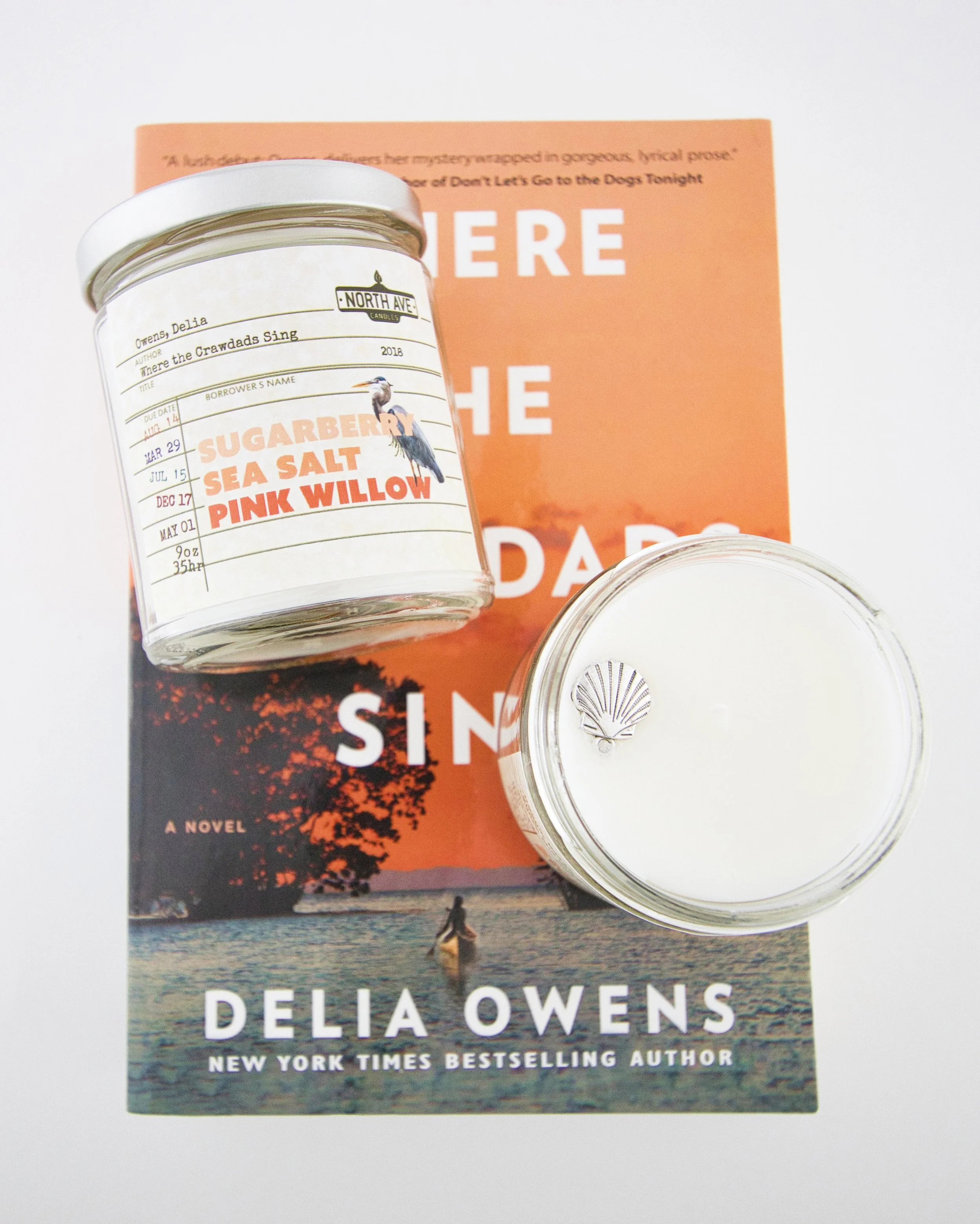 A book titled "Sing" by Delia Owens, a candle, and a jar of scented sea salt with labels reading "Sugarberry," "Sea Salt," and "Pink Willow" placed on top of the book.