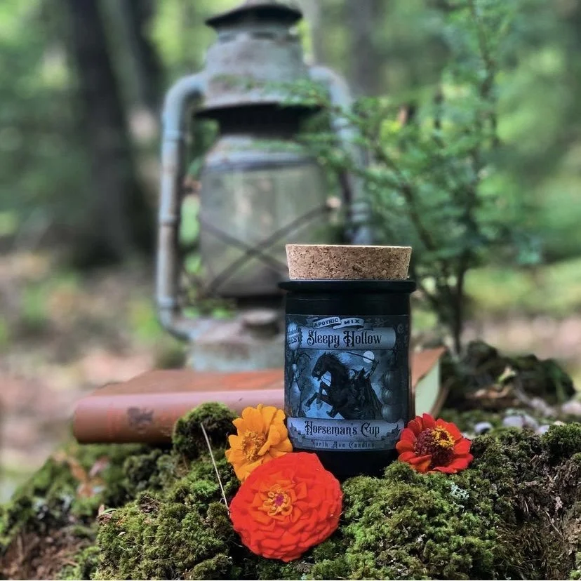 A black jar with a blue horse and moon design on it, labeled Sleepy Hollow Horseman's Cup, sits on mossy ground surrounded by vibrant orange and red flowers. Behind it, there is an old rusty lantern and a closed book in a forest setting.