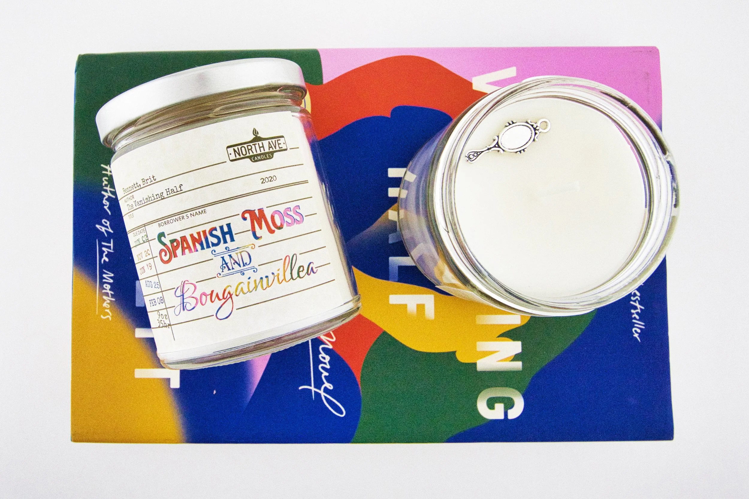 Two candles in glass containers on a colorful book cover. The label on one candle reads 'Spanish Moss and Bougainvillea,' with handwritten details. The book cover features bright, overlapping colors and white text.