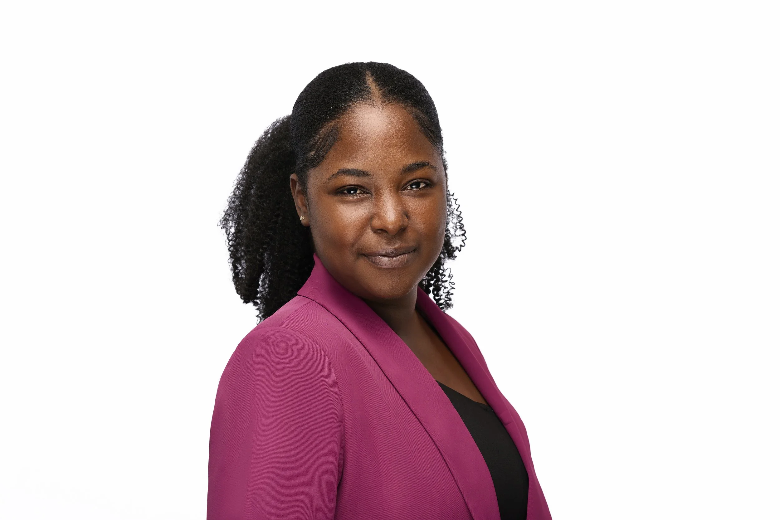 Portrait of a woman with dark curly hair tied back, wearing a pink blazer over a black top, standing against a white background