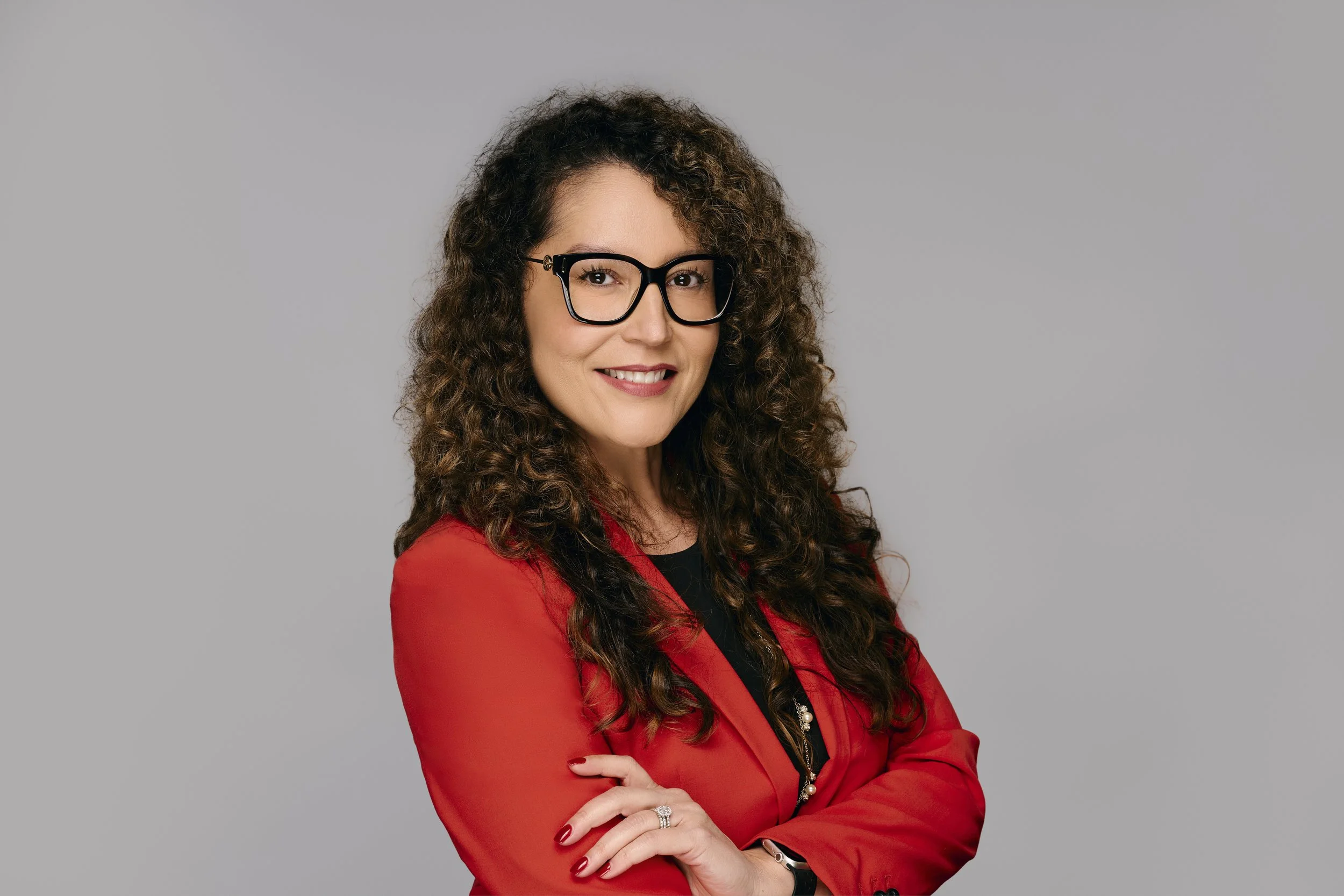 A woman with curly brown hair, wearing black glasses, a red blazer, and a black top, standing with her arms crossed against a plain gray background.