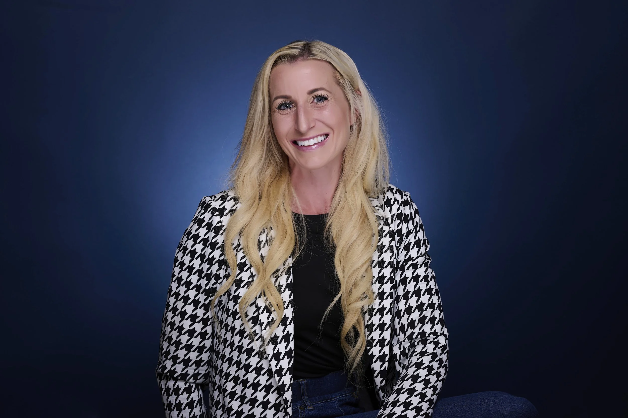 A woman with long blonde curly hair smiling, wearing a black and white houndstooth blazer and black top, against a dark blue background.