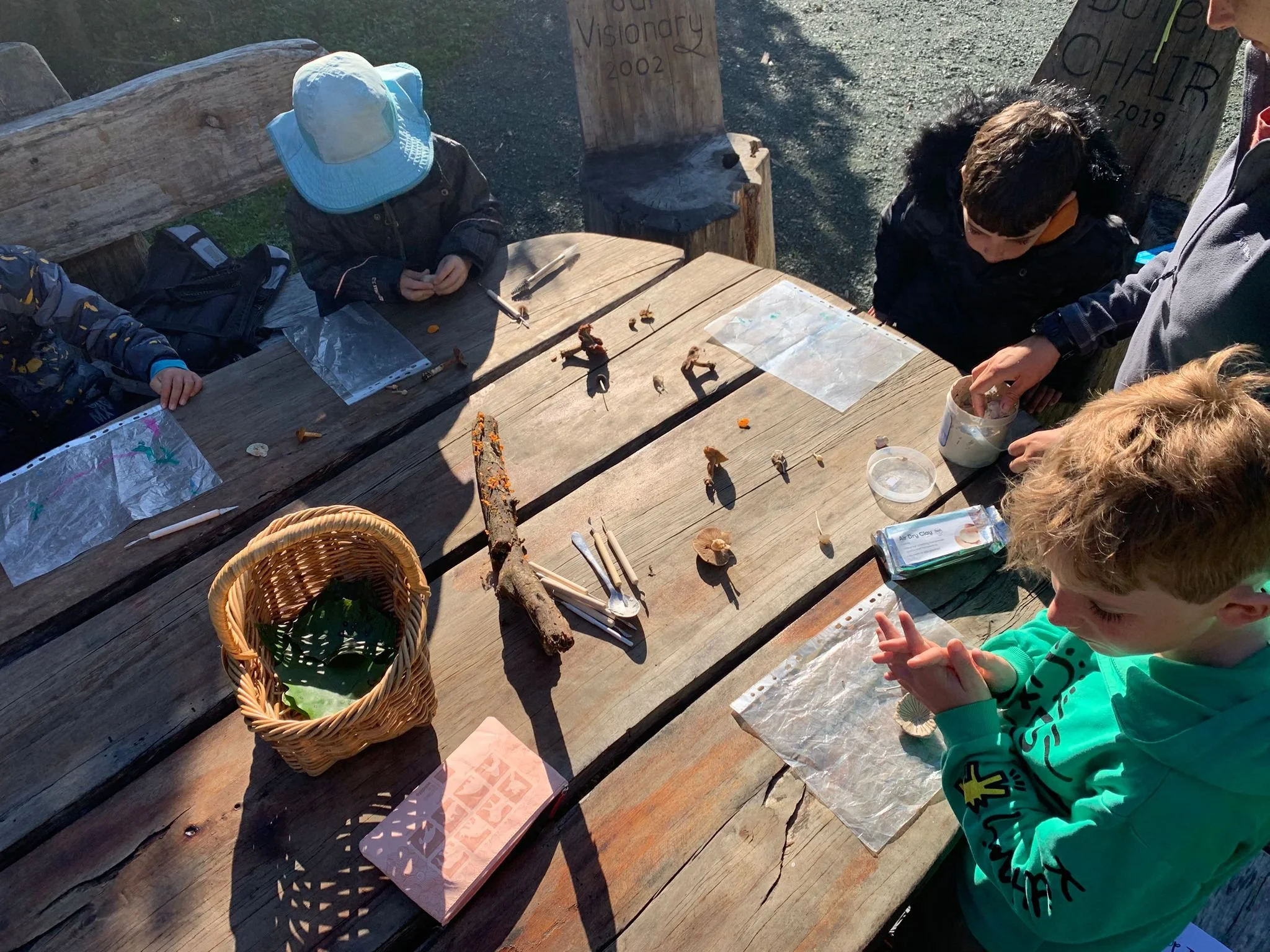 Children gathered around a wooden table outdoors, examining small natural objects and artifacts, with bags and plastic bags on the table, and a basket with leaves.