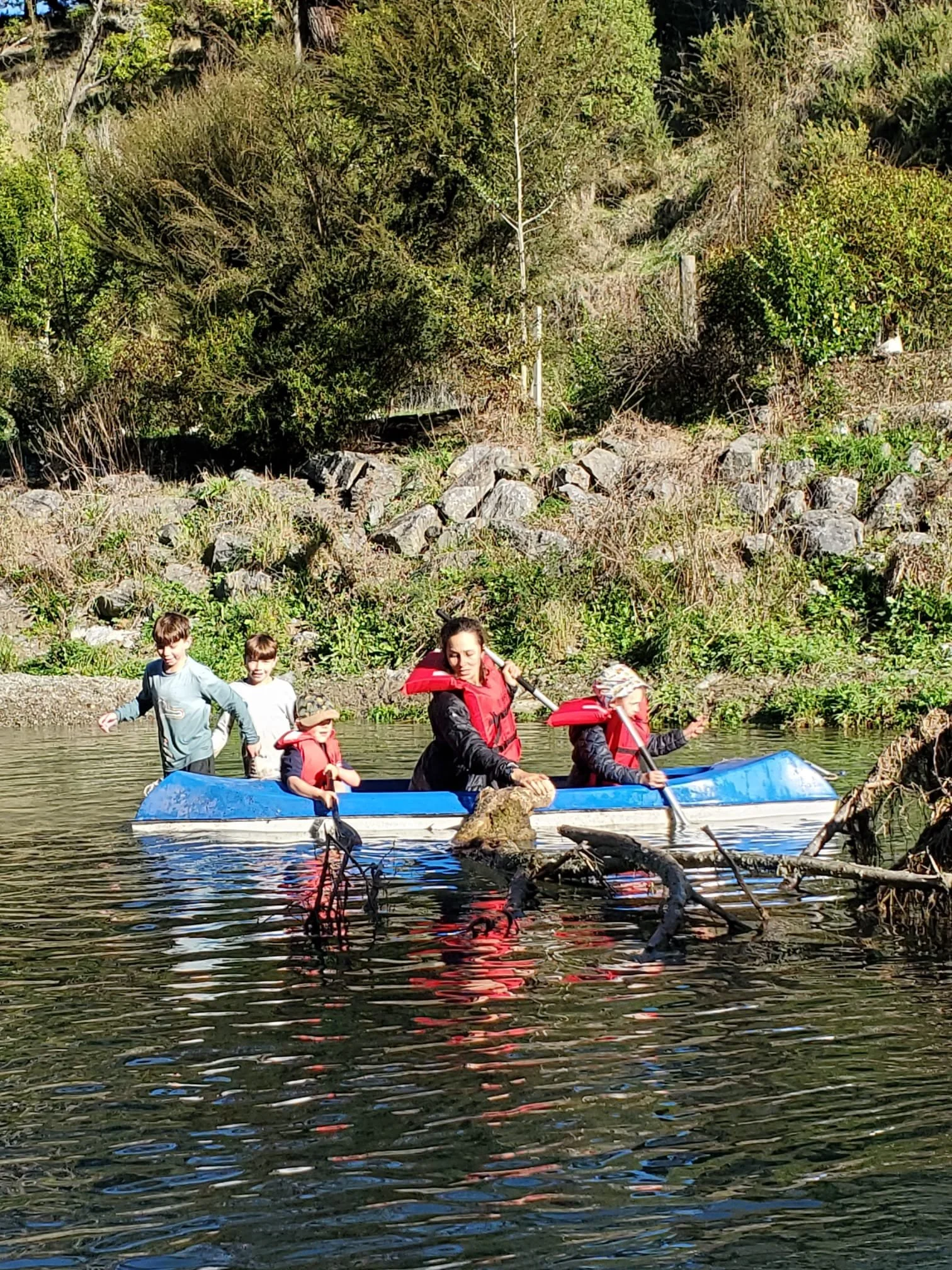 Group of children and a woman paddling a blue canoe on a river surrounded by green trees and bushes.