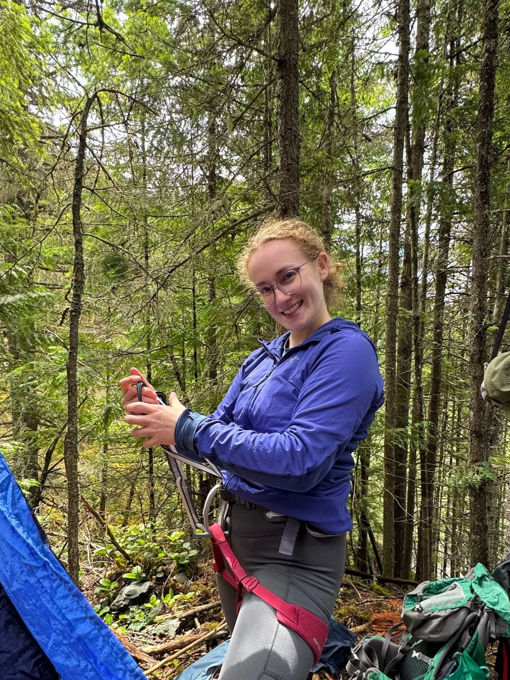 A woman with light brown curly hair, glasses, wearing a blue jacket, smiling and holding a small device, standing in a forest with tall trees and green foliage.