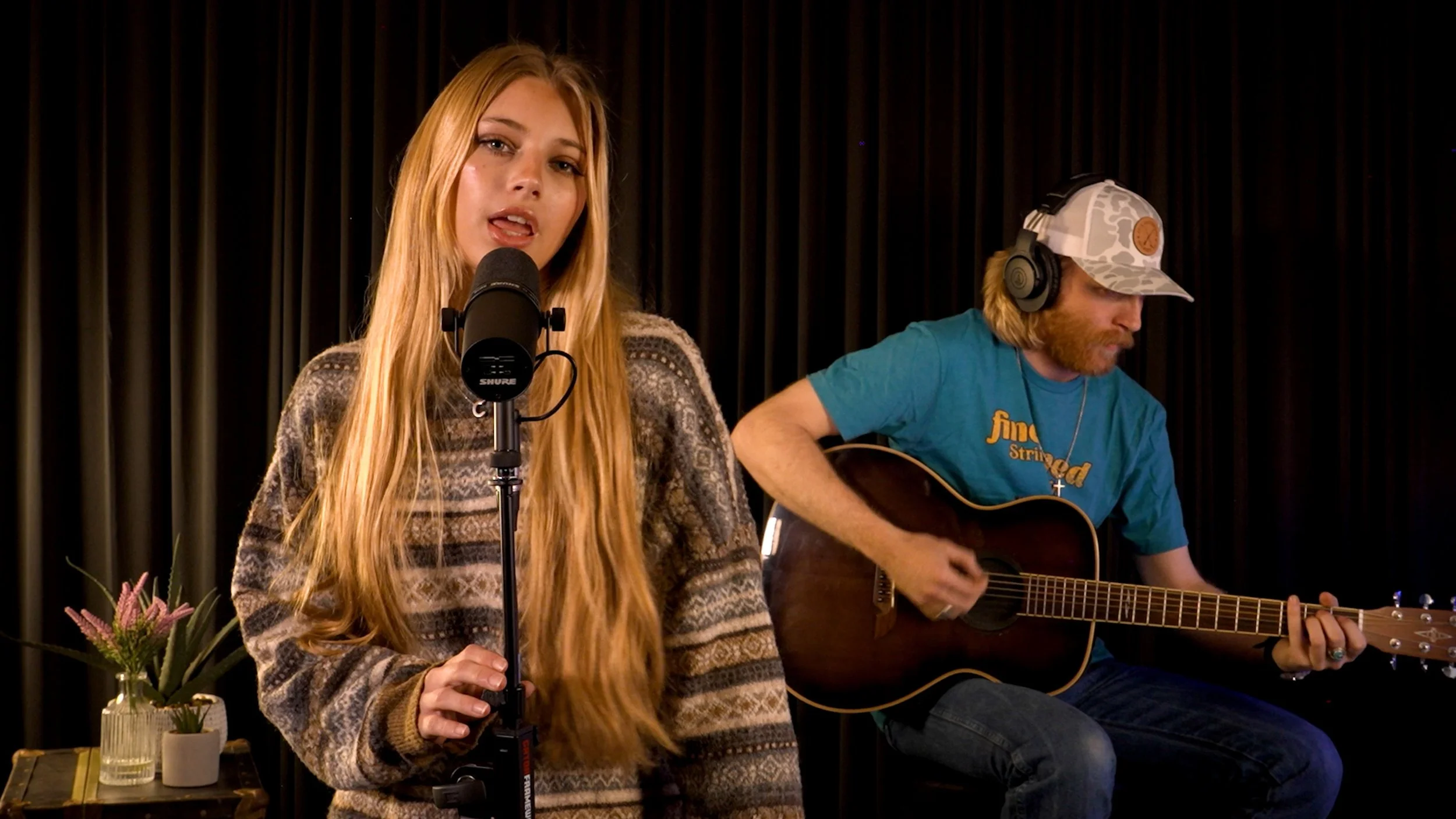 A woman singing into a microphone while a man plays an acoustic guitar beside her, with a dark curtain backdrop and flowers on a table in the foreground.