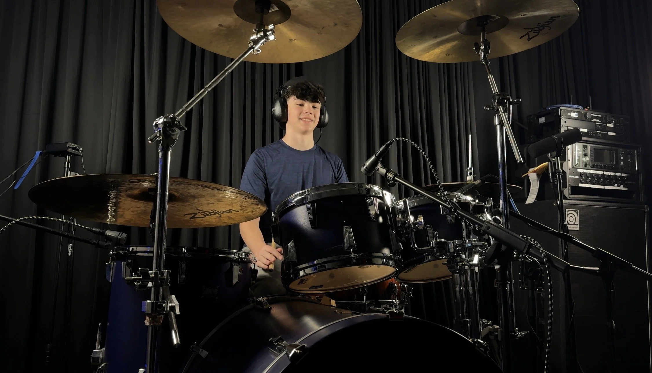 A young man wearing headphones playing a drum set in a music studio with black curtains and recording equipment.