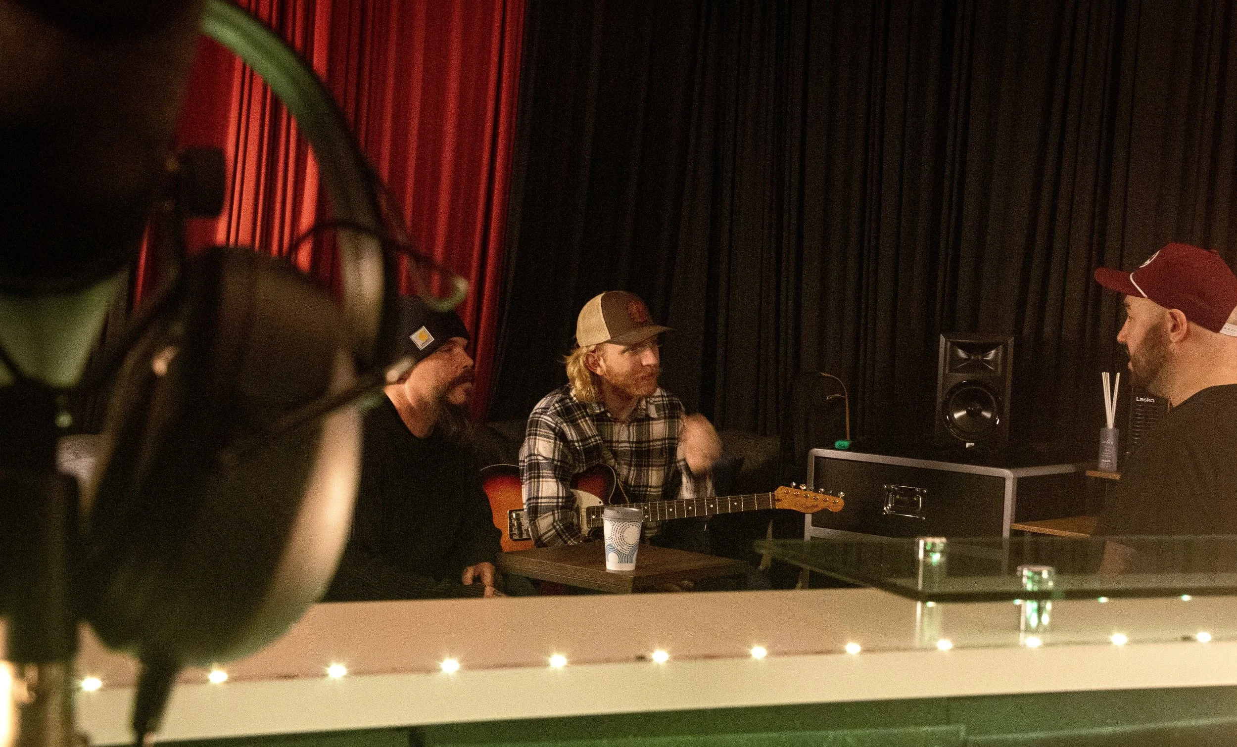 Three men sitting at a table in a recording studio, one playing an acoustic guitar, with dark curtains and a speaker in the background. One man wears a baseball cap, another a plaid shirt, and the third a maroon baseball cap, with a cup and electroni