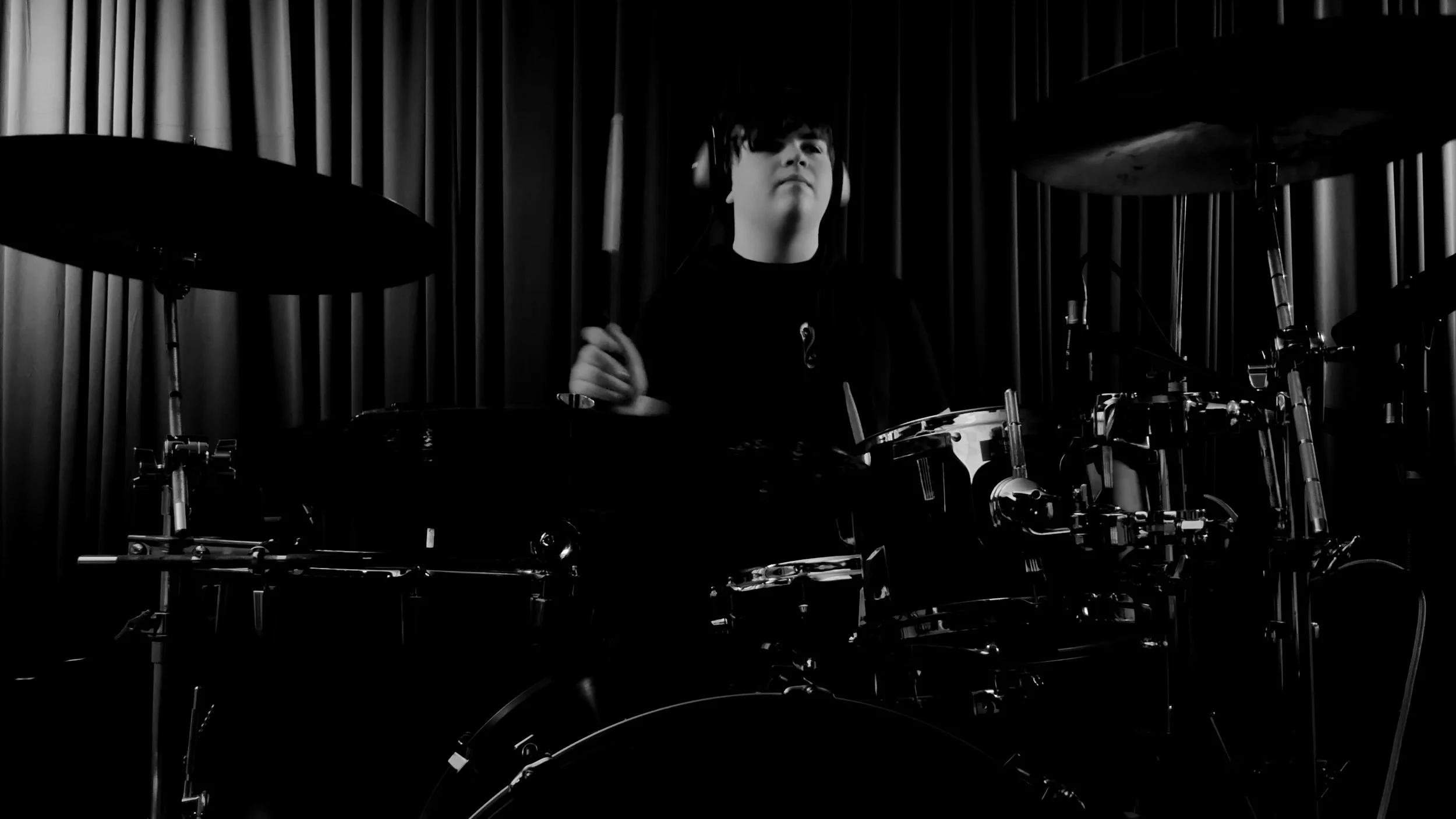 Black and white photo of a young man with headphones playing drums in a recording studio with vertical acoustic panels.