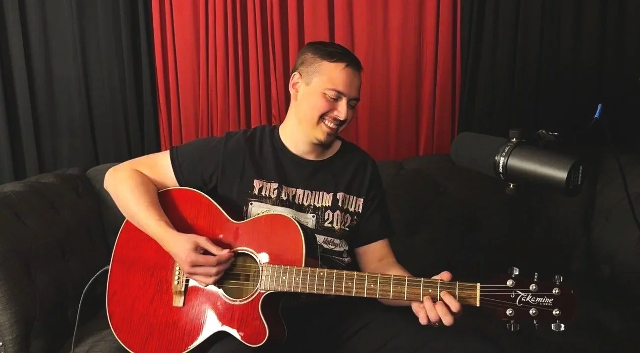 A man sitting on a black couch playing a red acoustic guitar, with a large microphone positioned nearby, in a room with black and red curtains.